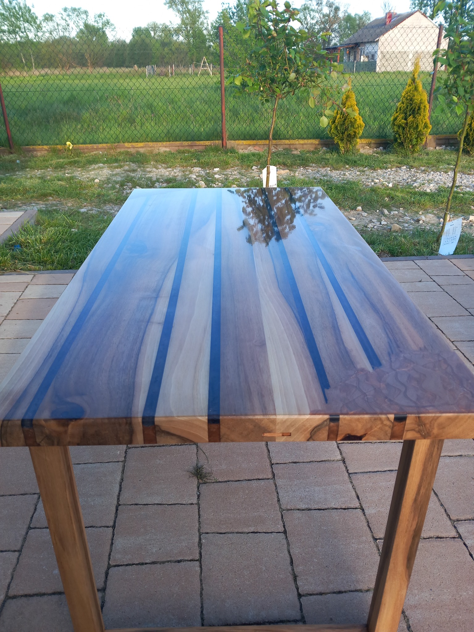 Stół z blatem z drewna egzotycznego z żywicą epoksydową, widoczne połączenia stolarskie, na zewnątrz, w tle trawa i drzewa.