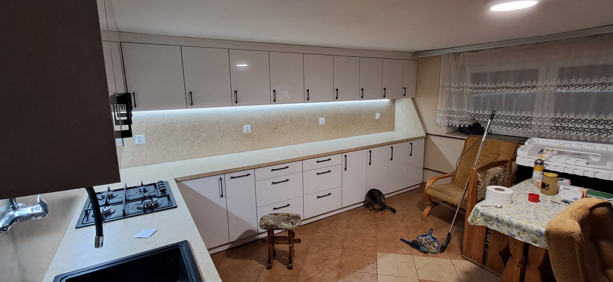 Jasna kuchnia w Nowym Korczynie z białymi szafkami z czarnymi uchwytami, podświetleniem LED pod szafkami wiszącymi i kotem siedzącym na podłodze.