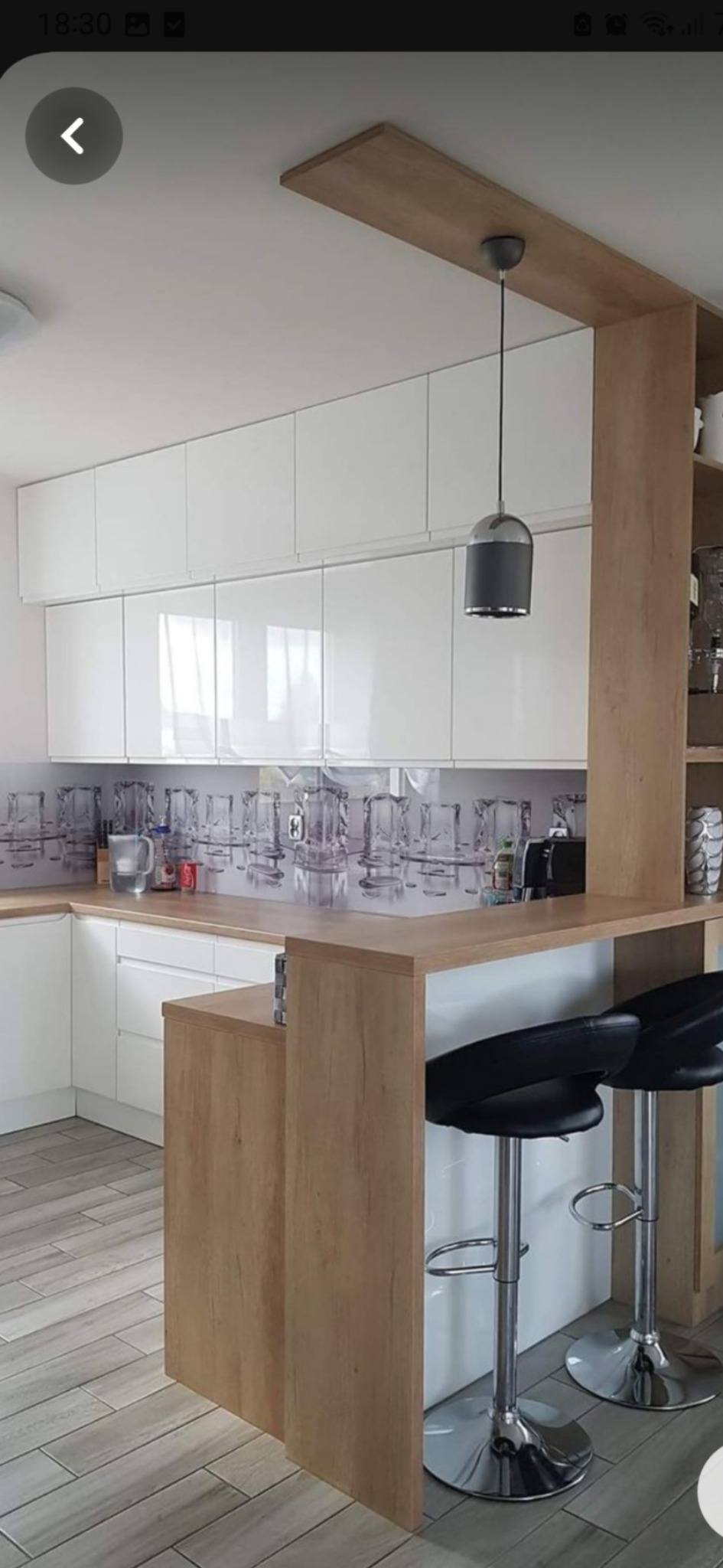 Biała kuchnia z błyszczącymi frontami, drewnianym blatem i barkiem oddzielającym przestrzeń, uzupełniona o dwa chromowane hokery z czarnymi siedziskami oraz szklany panel nad blatem z motywem...