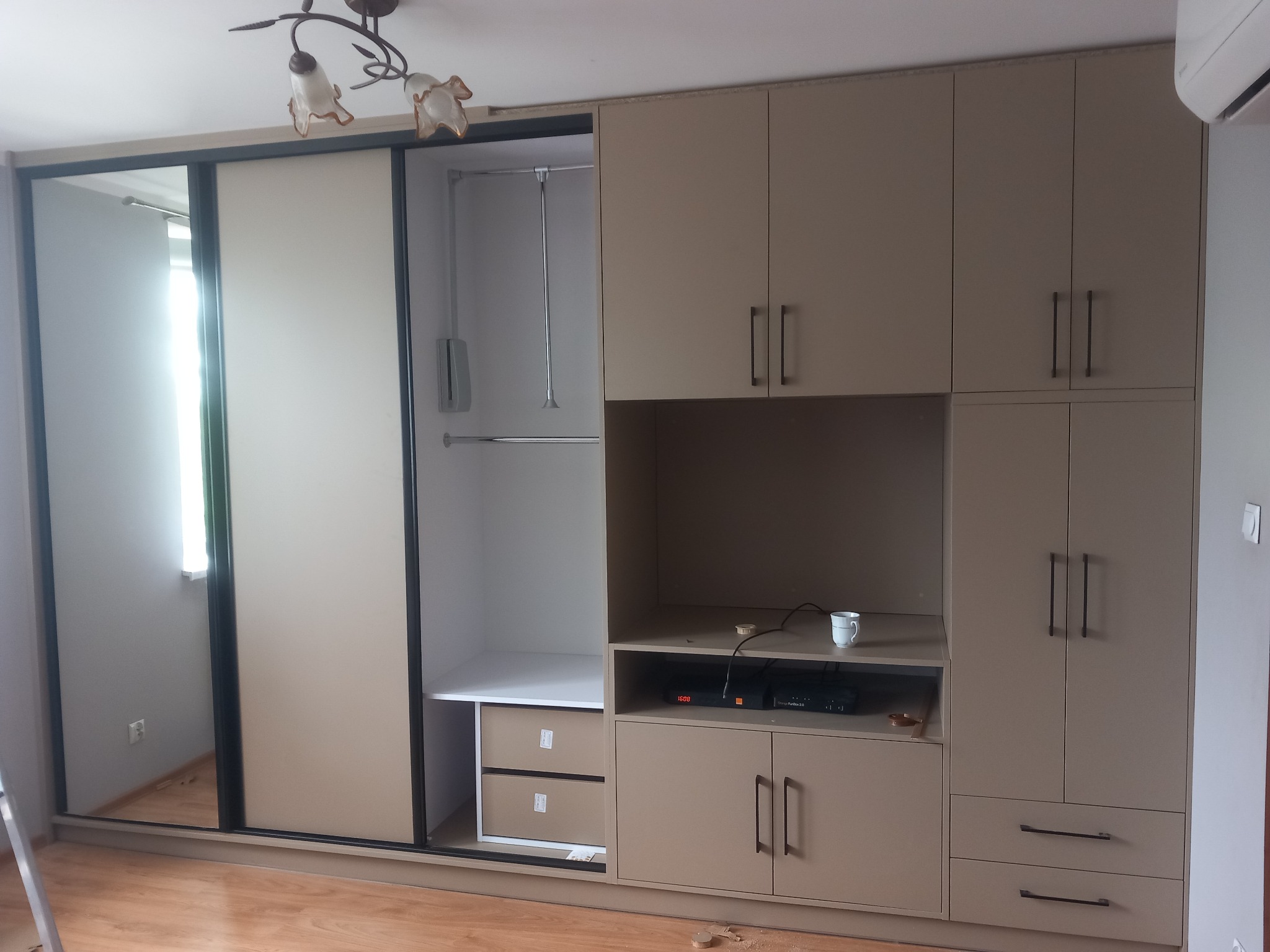 Beżowa szafa wnękowa z przesuwnymi drzwiami i częścią RTV, zlokalizowana w Nowym Korczynie, z widocznymi niedoskonałościami wykończenia i kubkiem na półce.