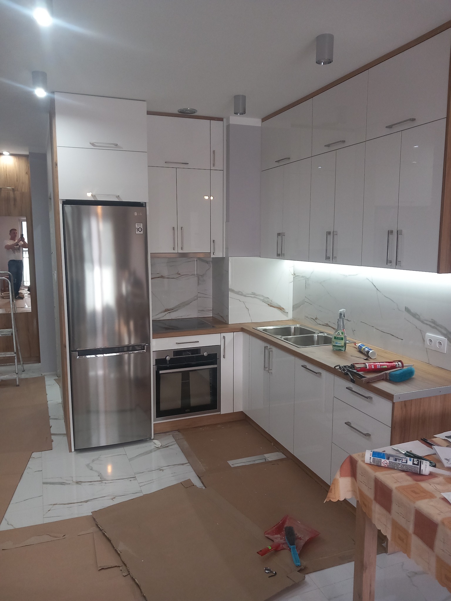 Nowoczesna, biała kuchnia z drewnianymi akcentami w trakcie montażu, widoczne narzędzia i zabezpieczenia podłogi kartonem, osoba w odbiciu lodówki robiąca zdjęcie.