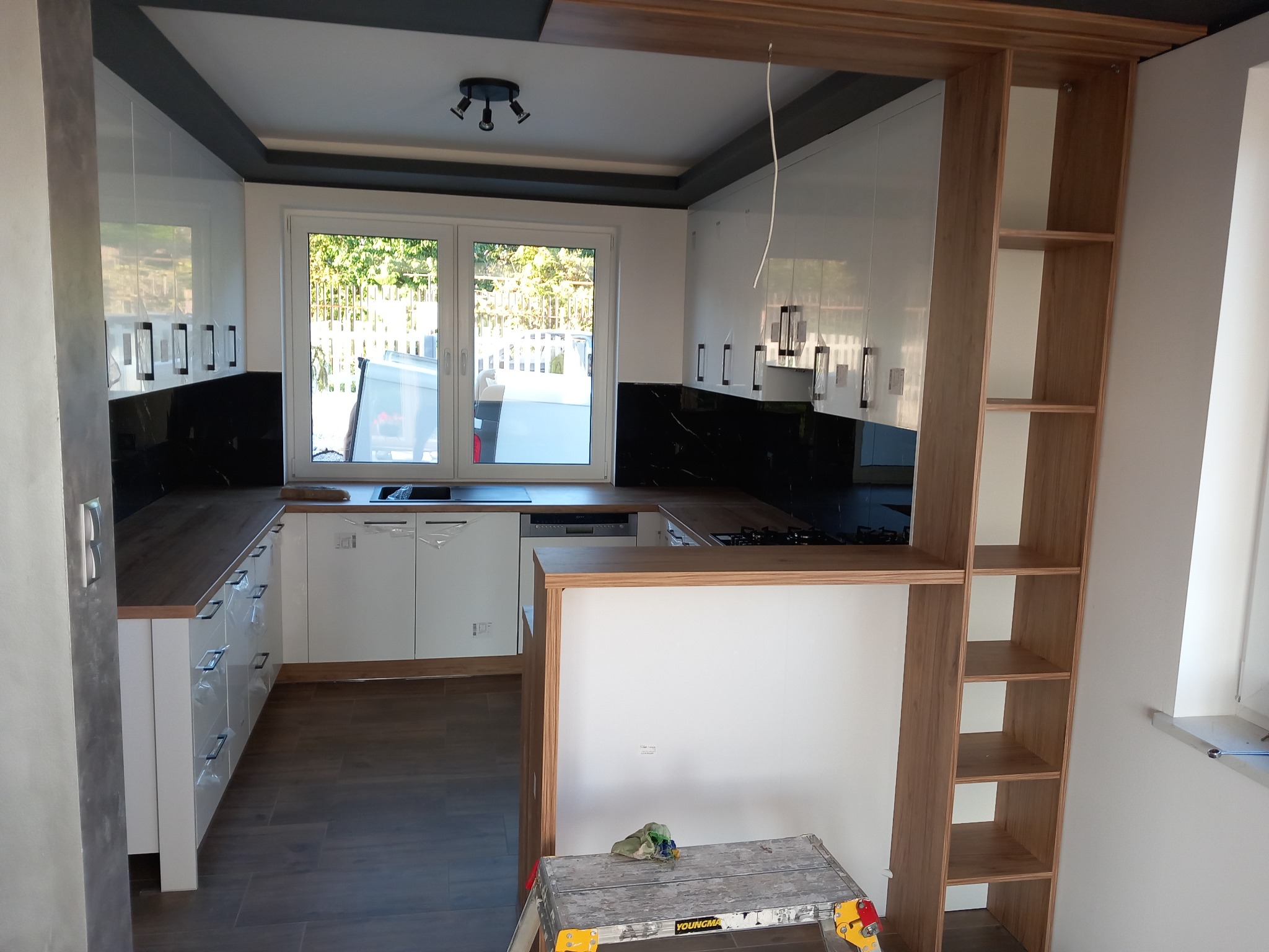 Nowoczesna kuchnia z białymi szafkami i czarnym backsplashiem, widoczna przez otwarte przejście z drewnianymi półkami, drabina malarska w dolnym rogu.