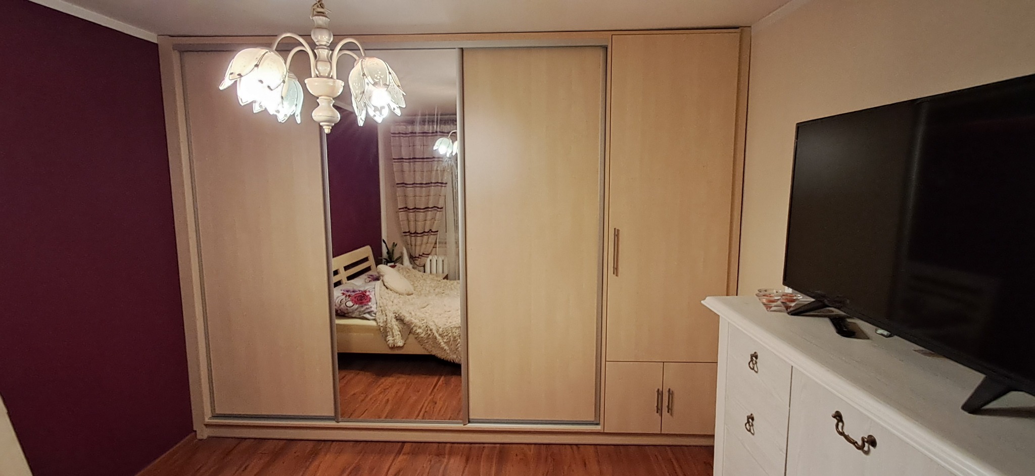 Beżowa szafa wnękowa z przesuwnymi drzwiami lustrzanymi, odbijająca wnętrze sypialni z łóżkiem i wzorzystymi zasłonami, oświetlona ozdobnym żyrandolem.