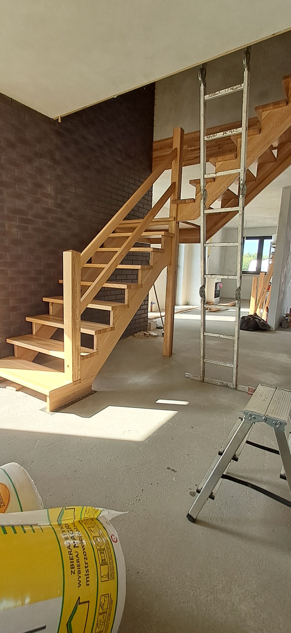 Jasne, drewniane schody z balustradą w surowym wnętrzu. Widoczna drabina i worki z materiałami budowlanymi. Ceglana ściana w tle. Etap prac wykończeniowych.