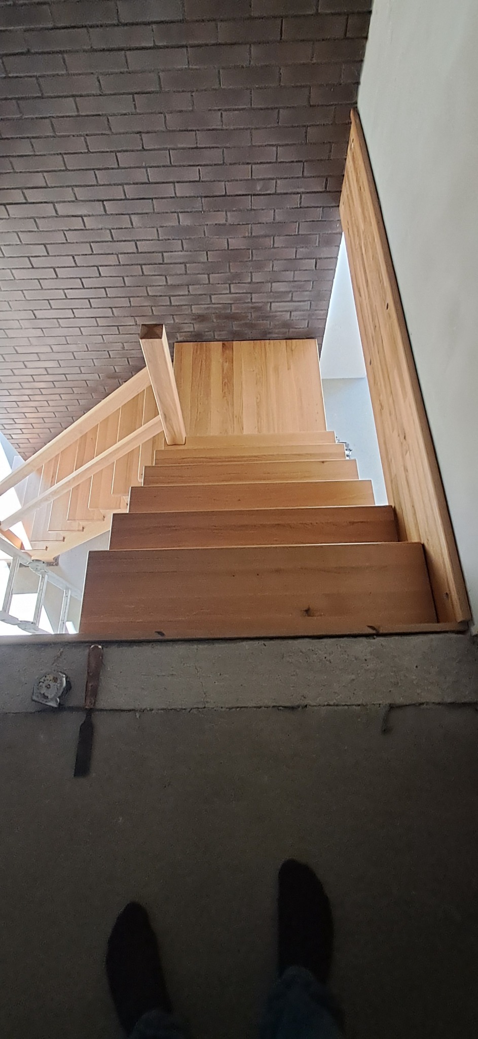 Wnętrze z widokiem na drewniane schody z barierką, prowadzące do góry. Na podłodze widoczne narzędzia i stopy osoby robiącej zdjęcie. Ceglana ściana w tle.