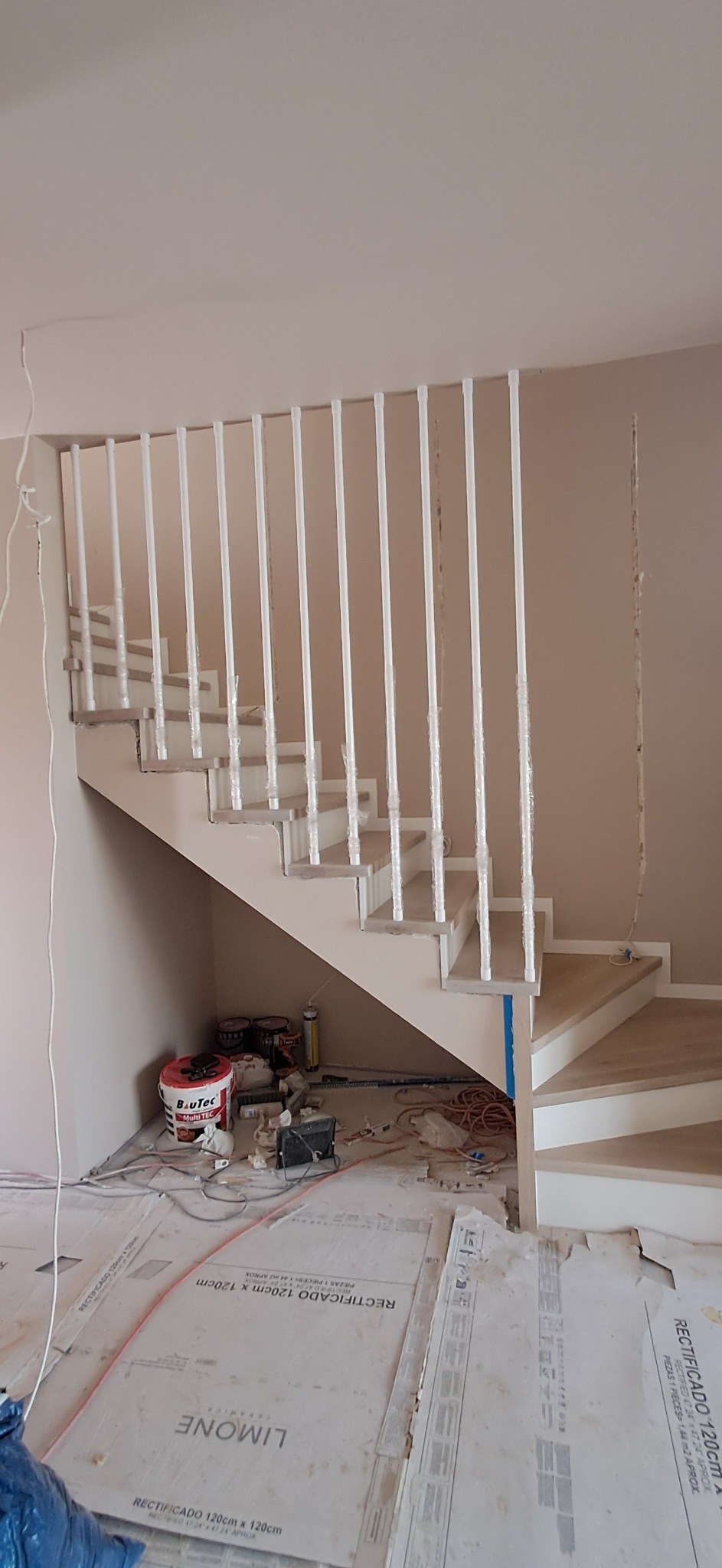 Montaż schodów drewnianych z białą balustradą w trakcie prac wykończeniowych. Podłoga zabezpieczona kartonami, widoczne narzędzia i puszki farb.