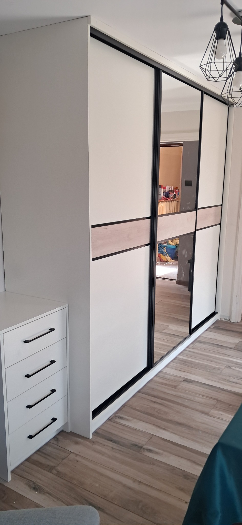 Biała szafa przesuwna z lustrem i drewnianym akcentem na środku, widoczna obok biała komoda z czarnymi uchwytami, na tle drewnopodobna podłoga.