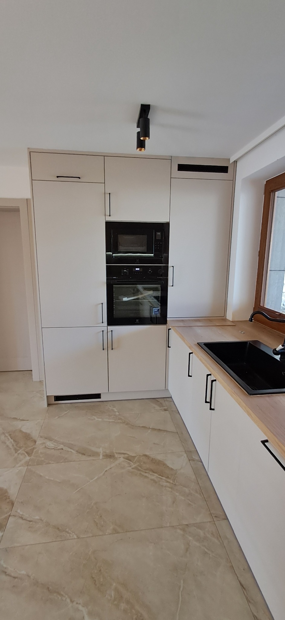 Jasna kuchnia na wymiar z zabudową piekarnika i mikrofalówki, czarny zlew i minimalistyczne uchwyty. Beżowe płytki podłogowe imitujące marmur.