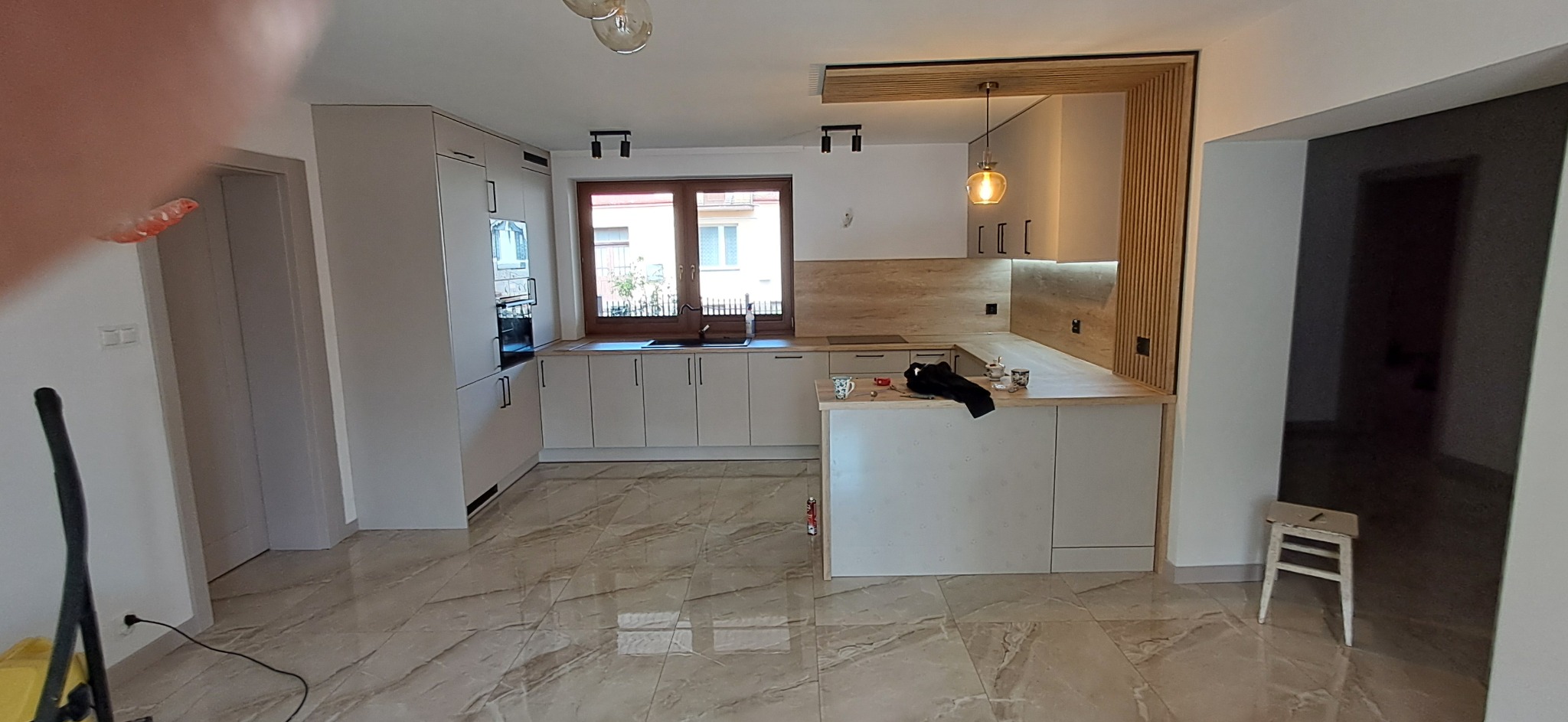 Jasna kuchnia z drewnianymi akcentami: zabudowa z półwyspem, wisząca lampa, okno z widokiem na ulicę, płytki podłogowe imitujące marmur. Nowoczesny design.