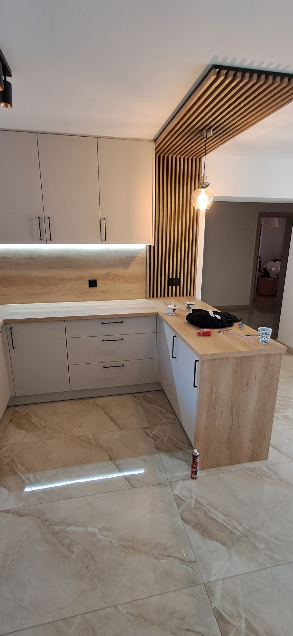 Jasna kuchnia na wymiar z drewnianymi akcentami: listwy sufitowe, blat i wykończenie wyspy. Nowoczesne oświetlenie i marmurowe płytki podłogowe.