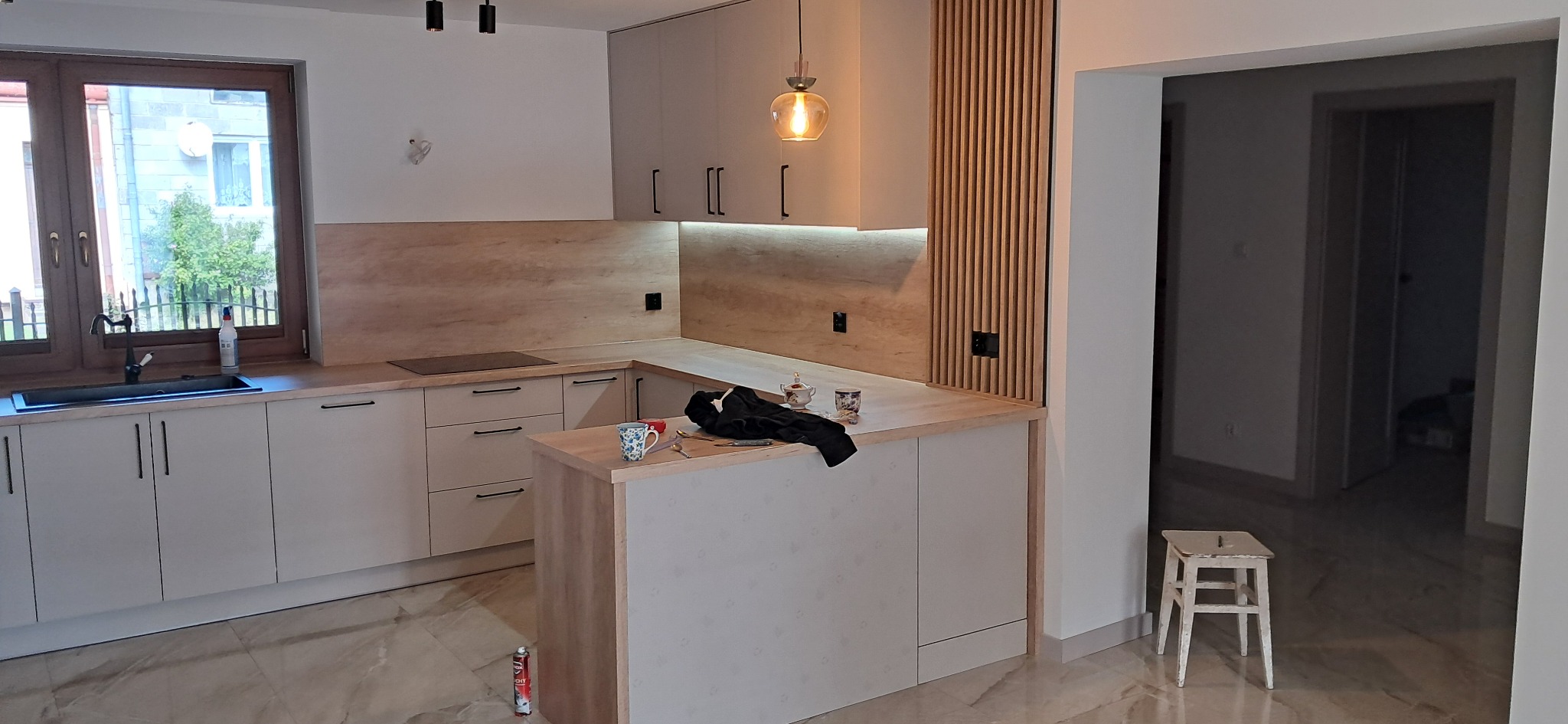 Jasna kuchnia z drewnianym akcentem, wyspą i oświetleniem punktowym, widok na okno i sąsiedni pokój, krzesło przy wejściu.