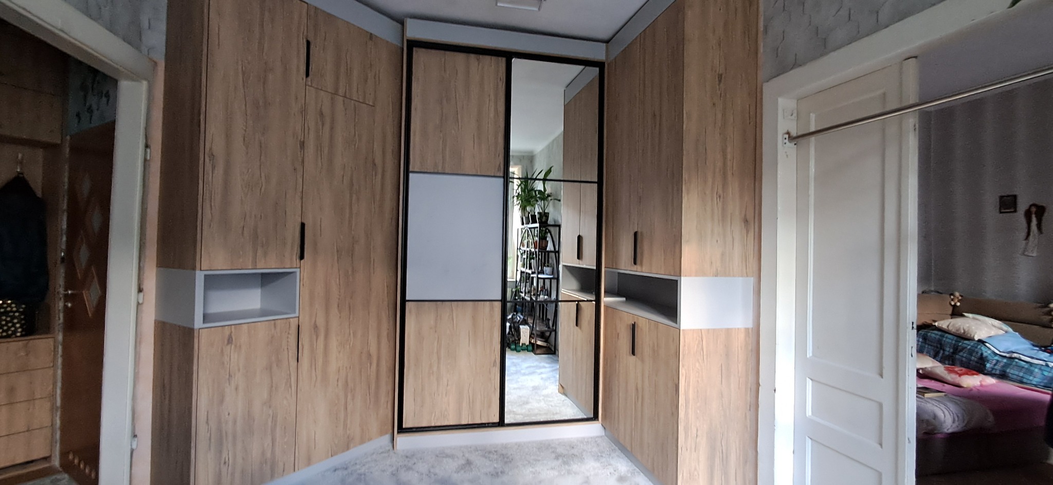 Zabudowa narożna z szaf z drewnopodobnymi frontami i lustrem w czarnej ramie, z otwartymi półkami. Widok z wnętrza pokoju na szafę i fragment drugiego pokoju.