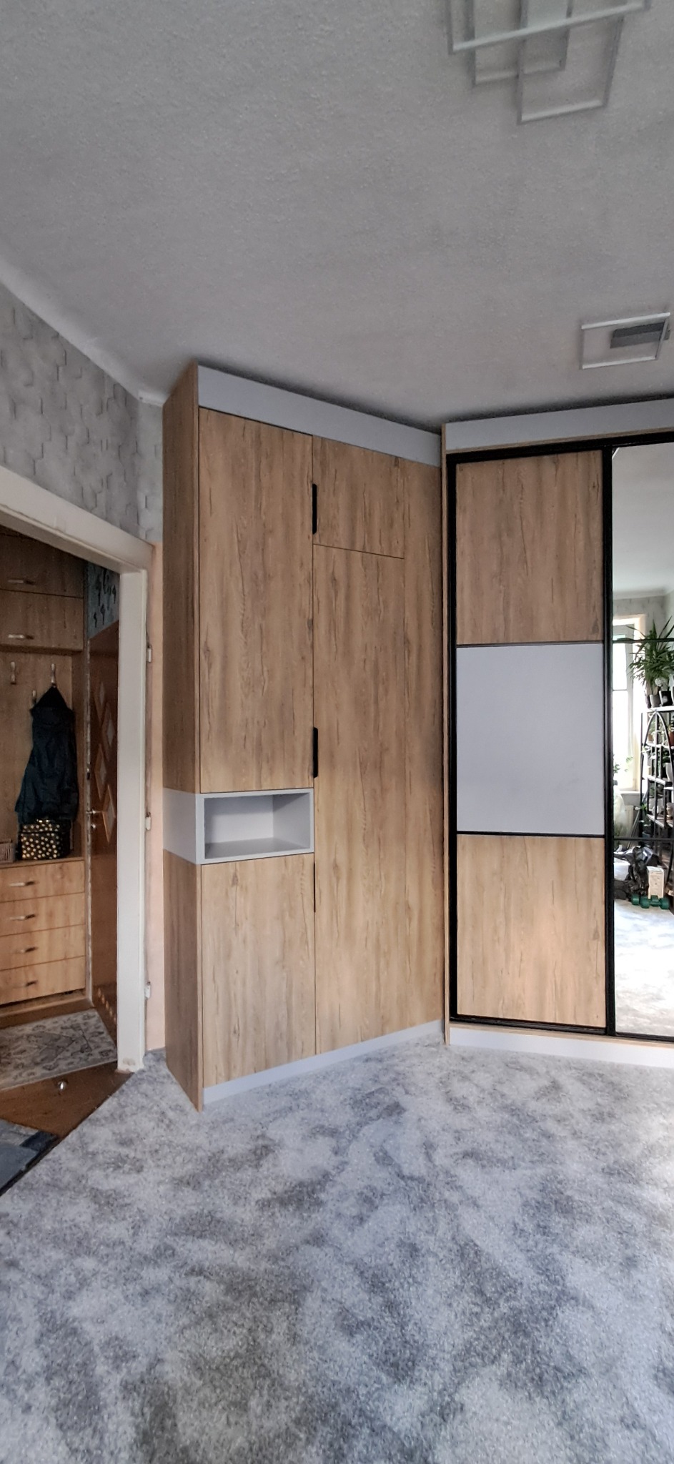 Funkcjonalna szafa wnękowa z drewnianym wykończeniem i lustrzanymi drzwiami przesuwnymi, uzupełniona otwartą półką, w jasnym wnętrzu pokoju.