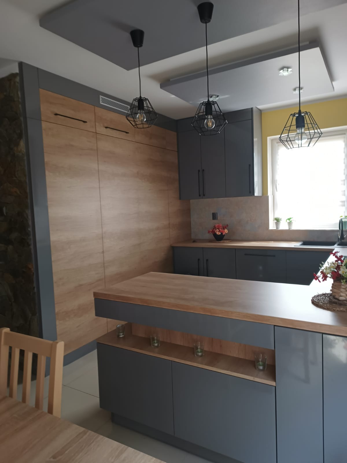 Szara kuchnia na wymiar z drewnianymi akcentami: wysoka zabudowa, wyspa z blatem, wiszące lampy w stylu industrialnym. Żółta ściana w tle. Nowoczesny design.