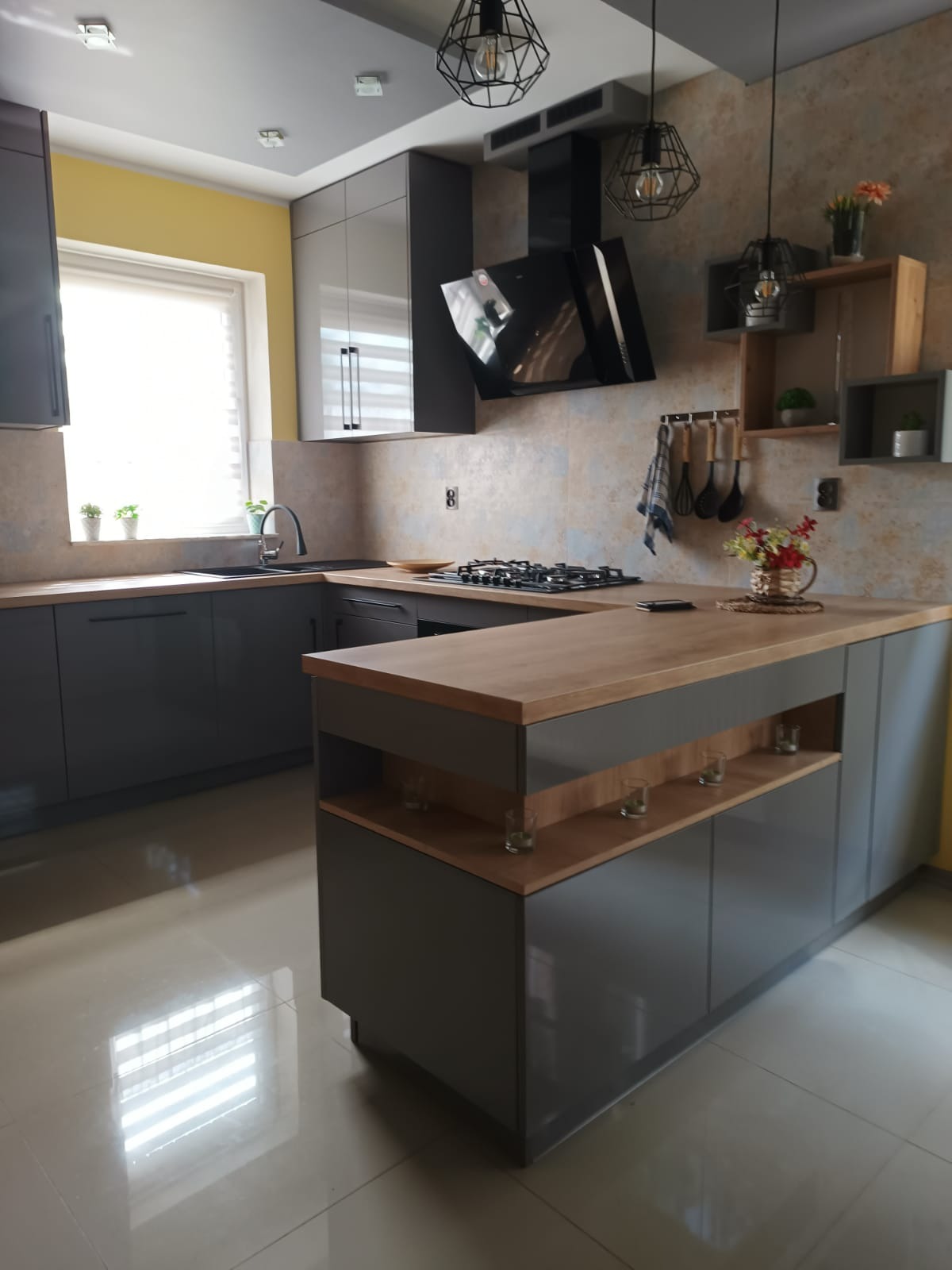 Nowoczesna kuchnia z szarymi szafkami, drewnianym blatem i wyspą. Oświetlenie punktowe i dekoracje ścienne dodają charakteru. Widok na okno z roletami.