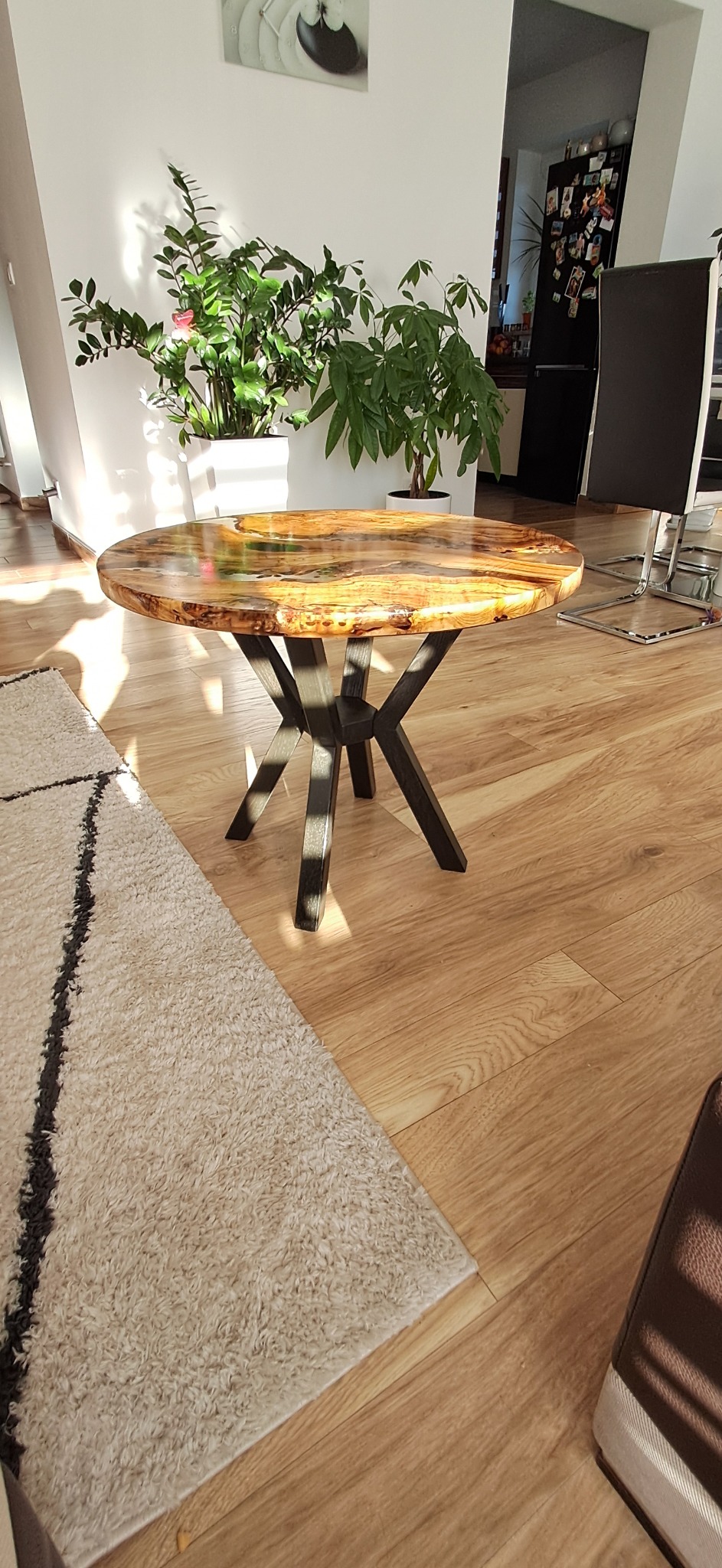 Okrągły stolik kawowy z blatem z żywicy epoksydowej i drewna, na metalowej podstawie w kształcie gwiazdy, w jasnym wnętrzu z drewnianą podłogą i roślinami doniczkowymi.