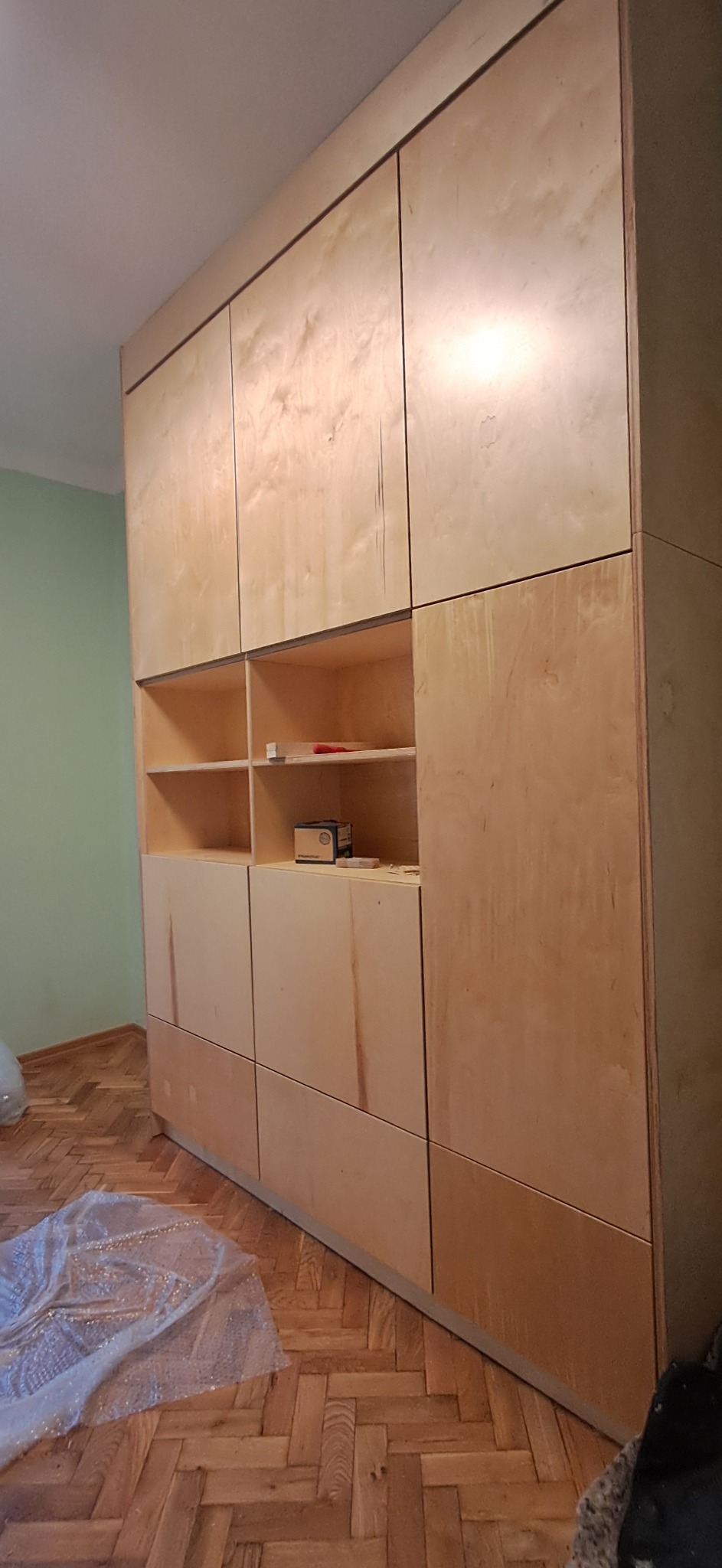 Duża, jasna szafa z półkami, wykonana z drewna, z widocznymi detalami łączeń. Na podłodze folia ochronna, w tle zielona ściana. Minimalistyczny design.