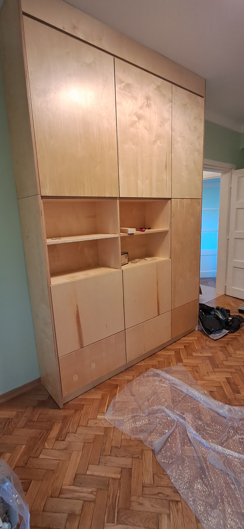 Wysoka, jasna szafa z surowej sklejki, częściowo z otwartymi półkami, stoi na parkiecie w jodełkę. Widoczne narzędzia i folia ochronna.