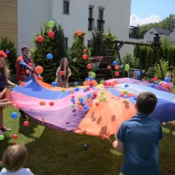 Grupa dzieci i dorosłych bawi się na trawie kolorową chustą animacyjną, podrzucając w górę kolorowe piłeczki.