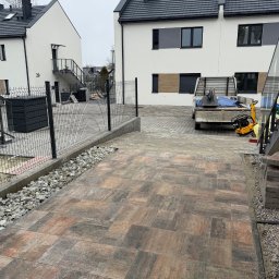 PRO HOUSE RAFAŁ SOBESTO - Ułożona kostka brukowa w odcieniach brązu i szarości na podjeździe przed nowoczesnymi domami. Widoczna ubijarka i materiały budowlane.