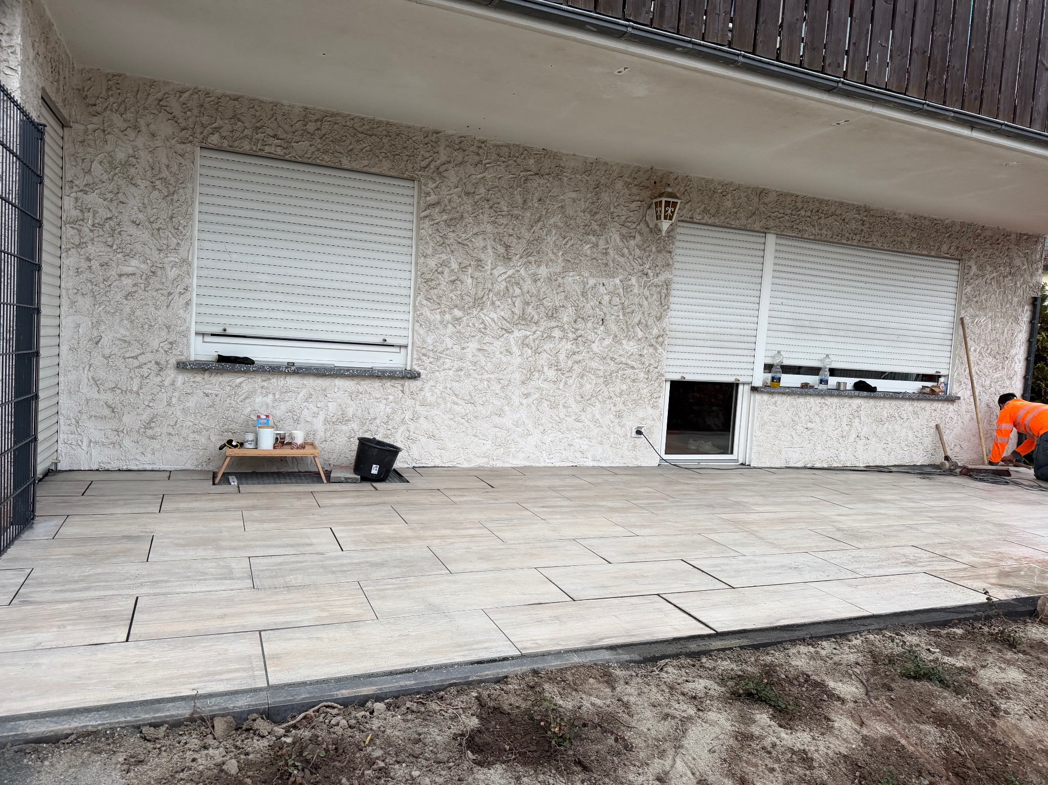 Jasne, prostokątne płyty tarasowe ułożone na zewnątrz budynku z fakturą. Widoczny pracownik w pomarańczowej kurtce i narzędzia. Częściowo odsłonięta ziemia.