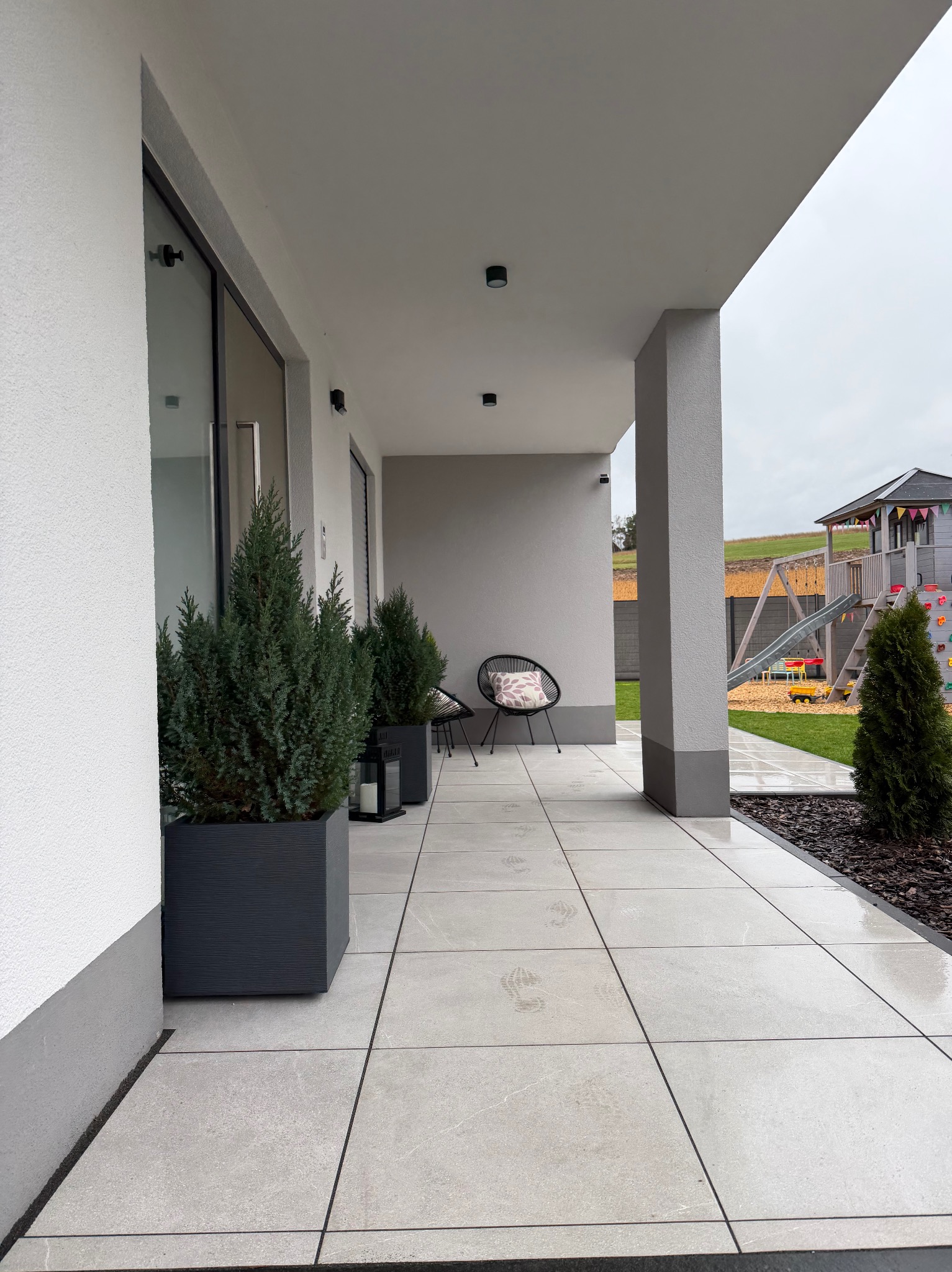 Nowoczesny taras z jasnych płyt, donice z iglakami, minimalistyczne krzesła. W tle plac zabaw i zielony trawnik. Widoczne ślady stóp na mokrej nawierzchni.