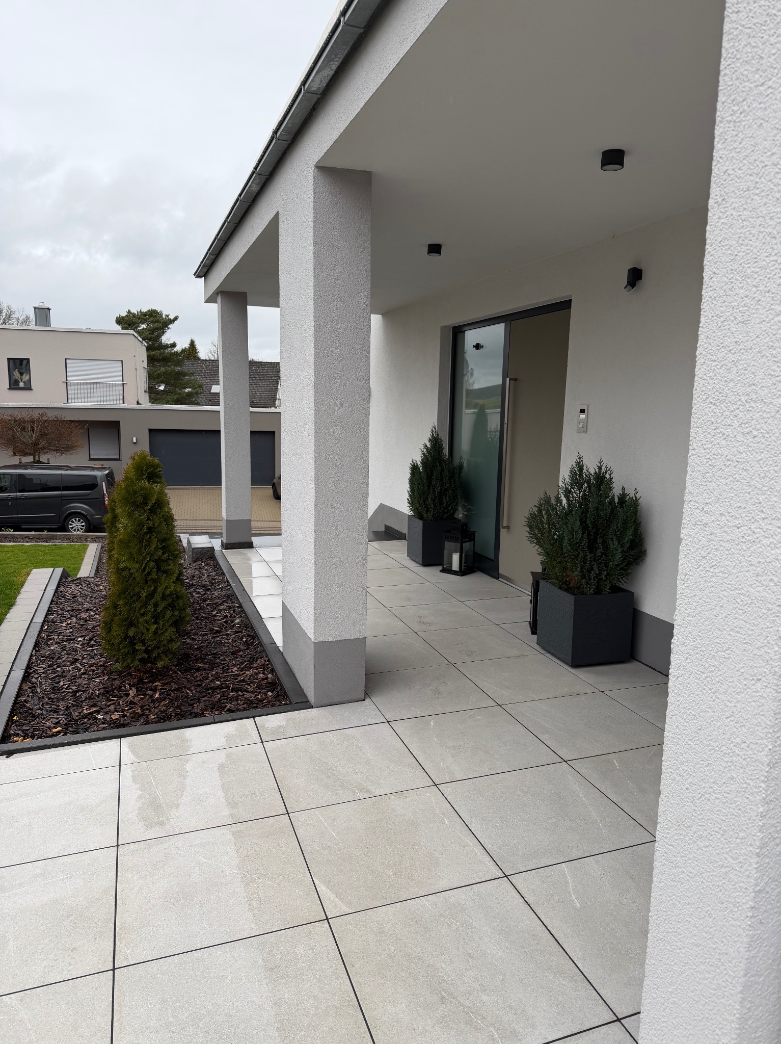 Nowoczesny taras z jasnoszarą kostką brukową, kolumnami i ozdobnymi roślinami w donicach. Minimalistyczny design z elementami zieleni.