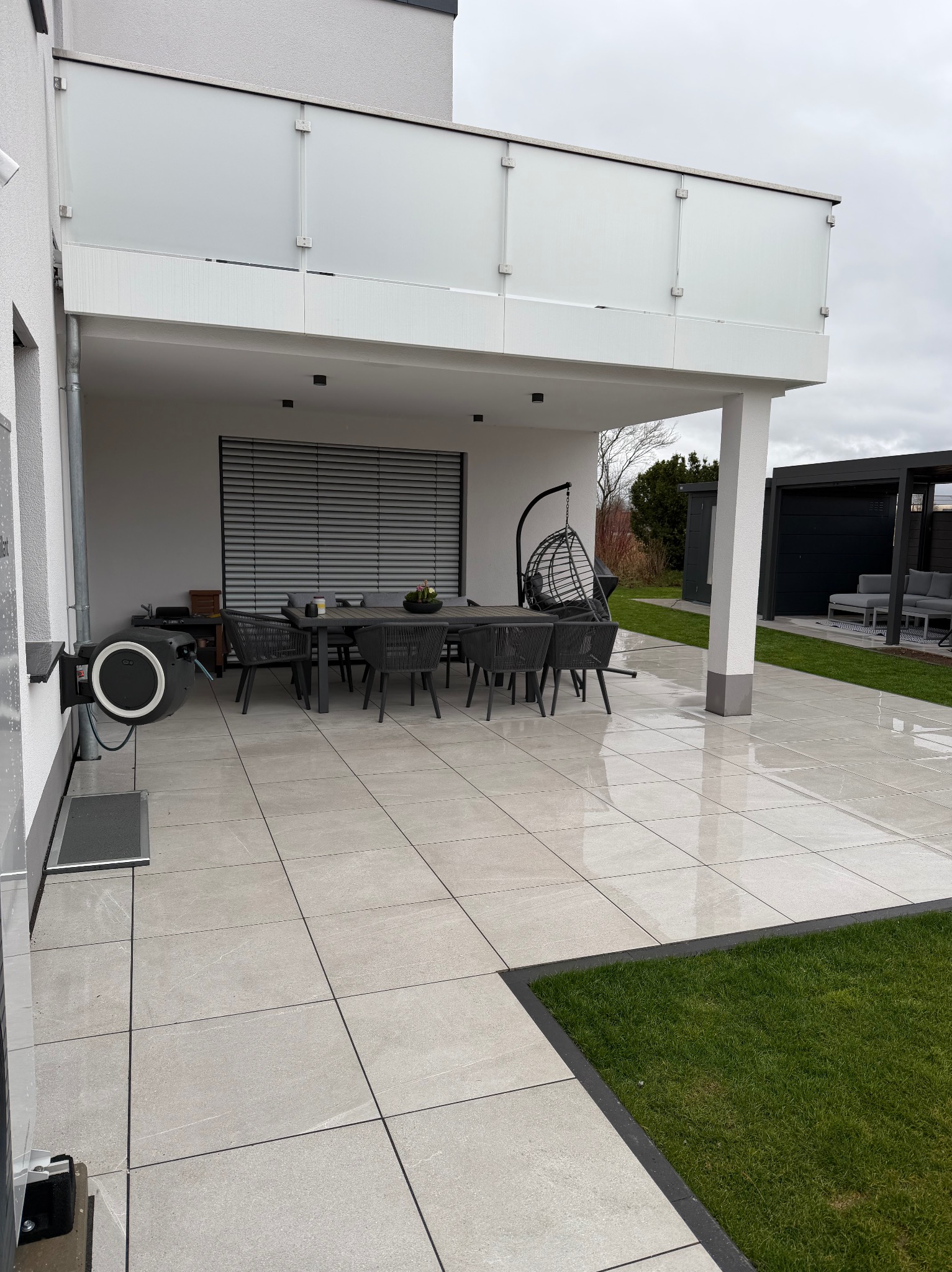 Nowoczesny taras z jasnych, kwadratowych płyt, ciemne obrzeże trawnika, meble ogrodowe i wiszące krzesło. Minimalistyczny design z elementami zieleni.