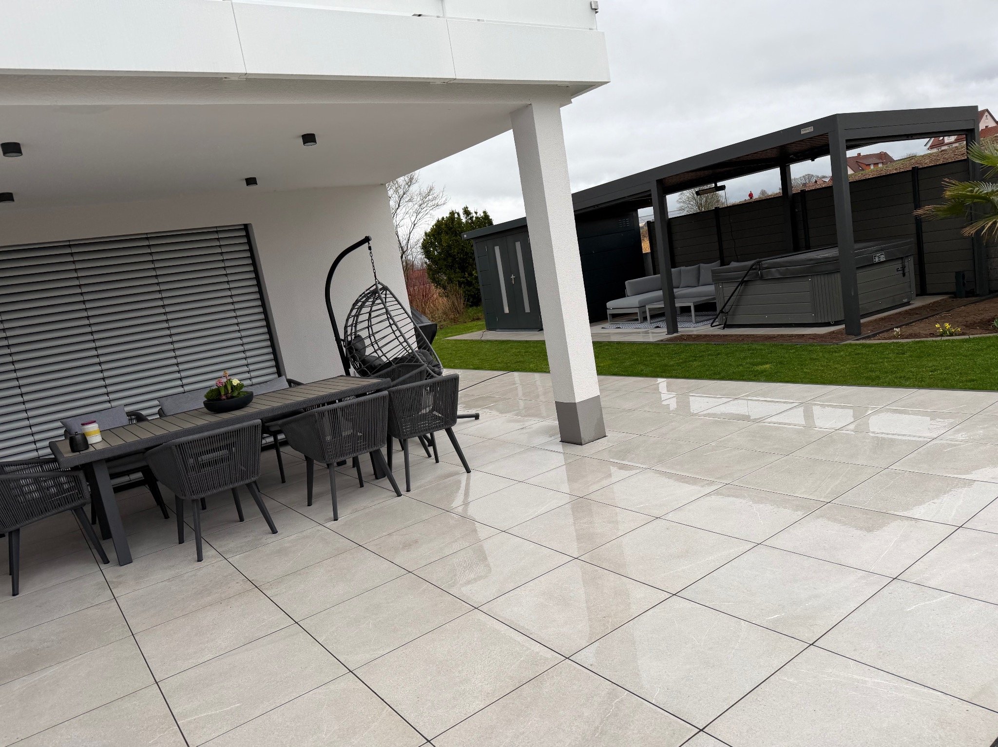 Nowoczesny taras z jasnych, kwadratowych płyt. Widoczny stół z krzesłami, huśtawka, pergola z jacuzzi i zadbany trawnik. Minimalistyczny design, stonowana kolorystyka.