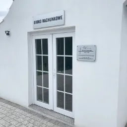 Biały budynek biura rachunkowego z dwuskrzydłowymi drzwiami wejściowymi, tabliczką informacyjną z nazwą firmy i danymi kontaktowymi, oświetleniem zewnętrznym oraz brukowanym chodnikiem.