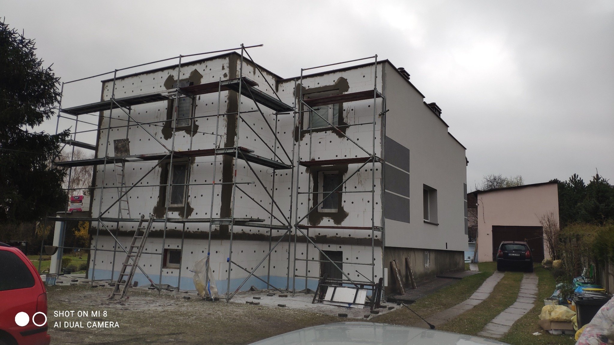 Dwupiętrowy dom w trakcie prac elewacyjnych, z widocznym rusztowaniem, częściowo ocieplony styropianem z charakterystycznymi okrągłymi wgłębieniami, fragment ściany już wykończony, pochmurne niebo...