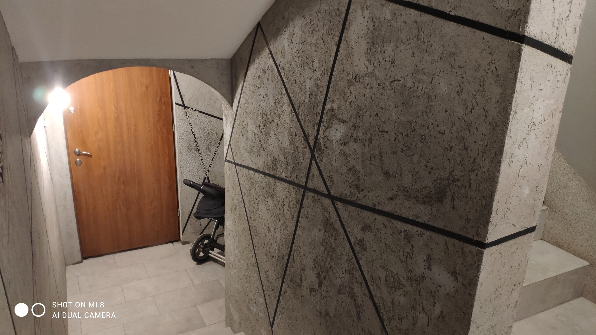 Wnętrze korytarza z dekoracyjną ścianą o fakturze betonu i geometrycznym wzorze z czarnych pasów, widoczna drewniana ościeżnica drzwi i fragment wózka dziecięcego.