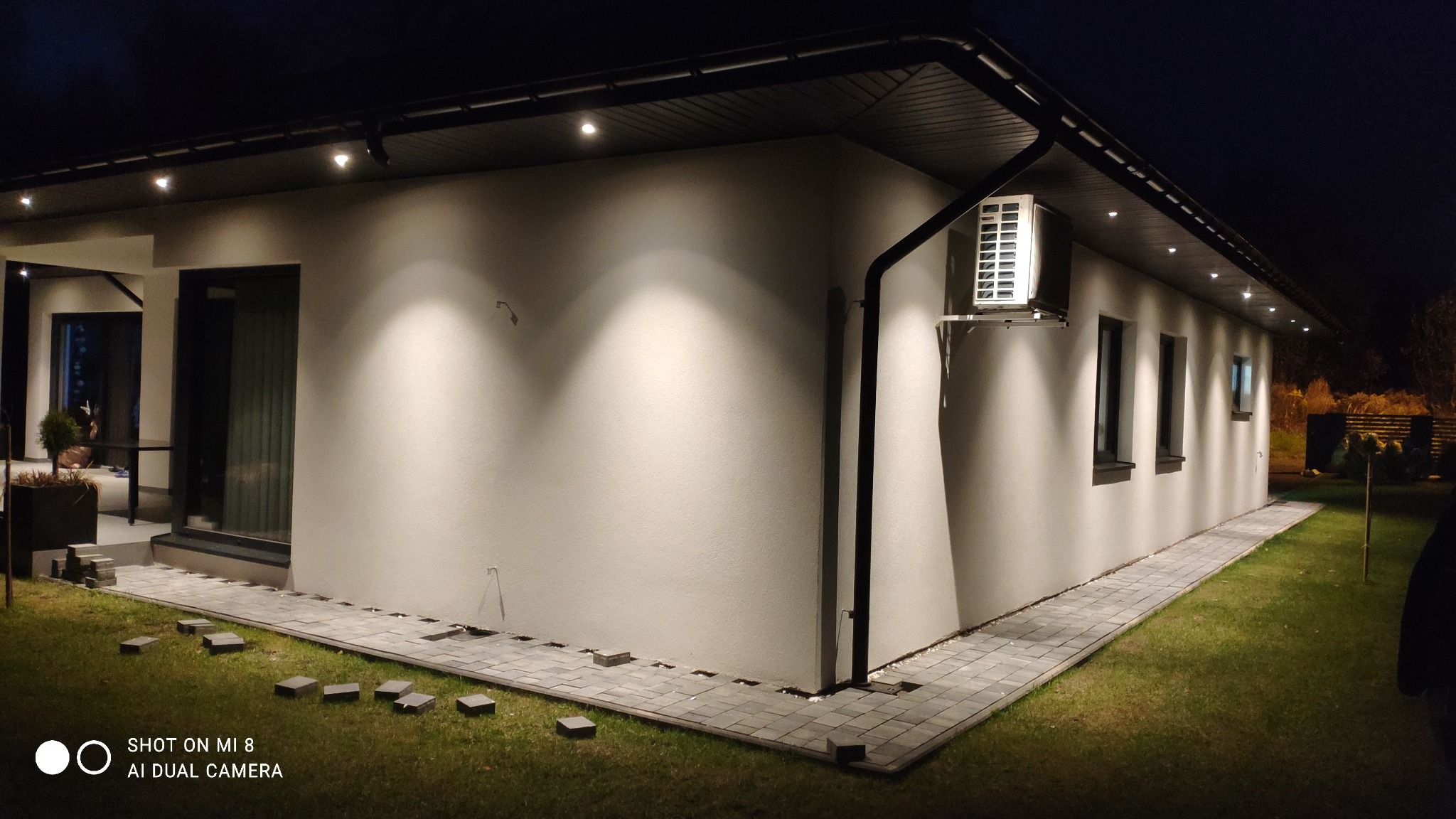 Nowoczesny dom jednorodzinny z szarą elewacją oświetloną punktowymi lampami wpuszczonymi w podbitkę, z widocznym klimatyzatorem na ścianie i chodnikiem z kostki brukowej wzdłuż elewacji...