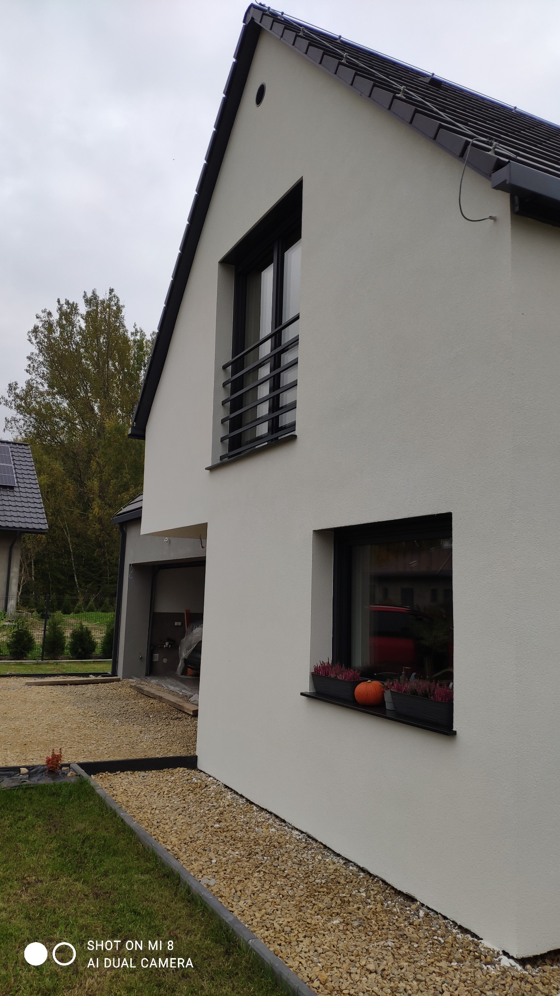 Nowoczesny dom jednorodzinny z jasną elewacją i czarnymi ramami okiennymi, widoczne kwiaty w donicach na parapecie i żwirowe podłoże.