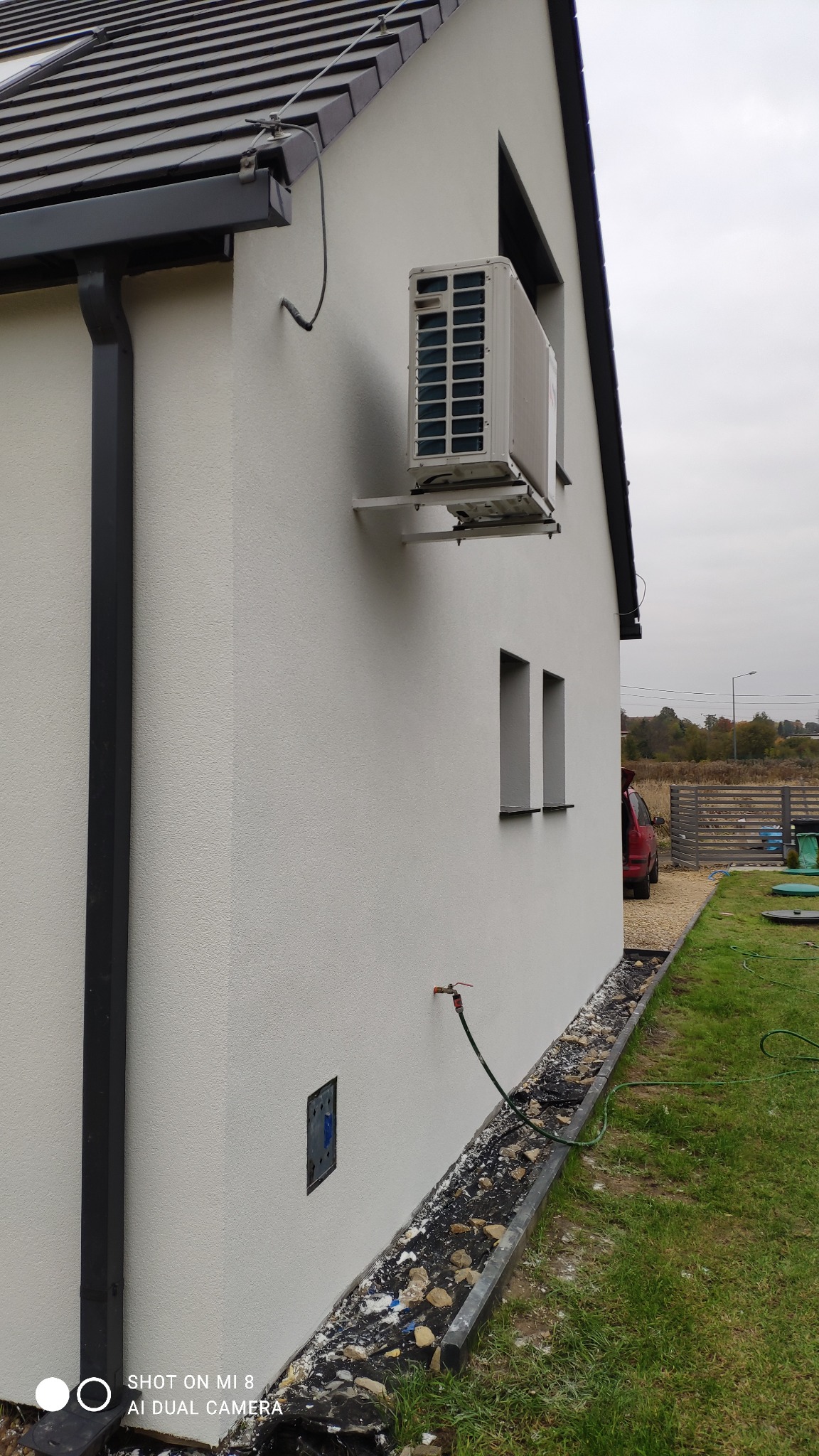 Boczny widok domu z jasną elewacją, widoczny zamontowany klimatyzator, rynna i fragment ogrodu z trawnikiem.