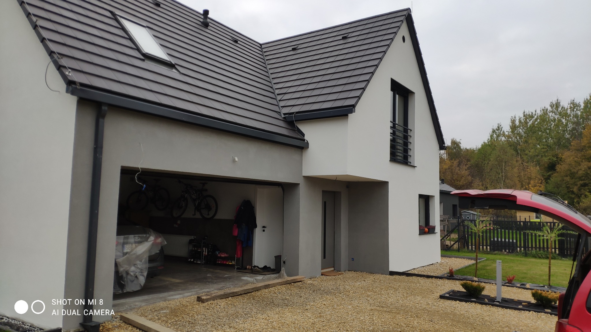 Nowoczesny dom jednorodzinny z garażem w trakcie wykańczania elewacji, widoczny szary tynk na części budynku i biała elewacja na pozostałej części, z czarną dachówką i elementami wykończeniowymi...