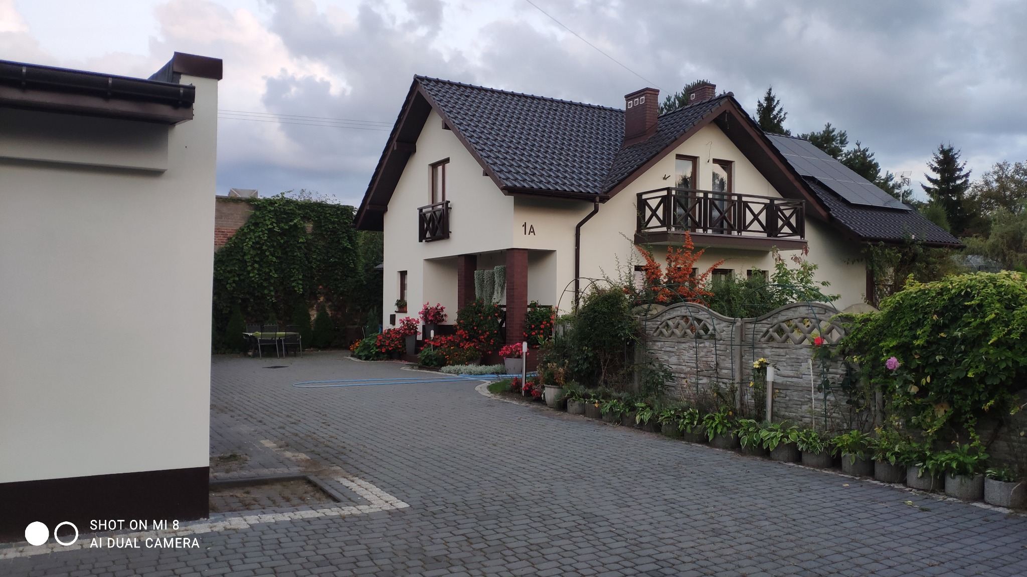 Dwupiętrowy dom z jasną elewacją, ciemną dachówką i panelami słonecznymi na dachu, widoczny z brukowanego podjazdu z dekoracyjną roślinnością i ogrodzeniem.