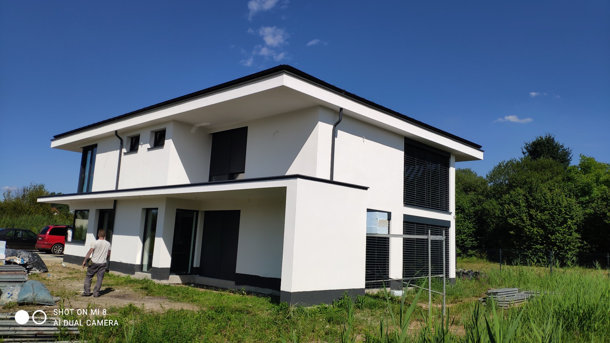 Nowoczesny, biały dom z płaskim dachem i czarnymi detalami elewacji, widok z zewnątrz, trawa i częściowo ułożone płyty chodnikowe na pierwszym planie, pracownik w tle.