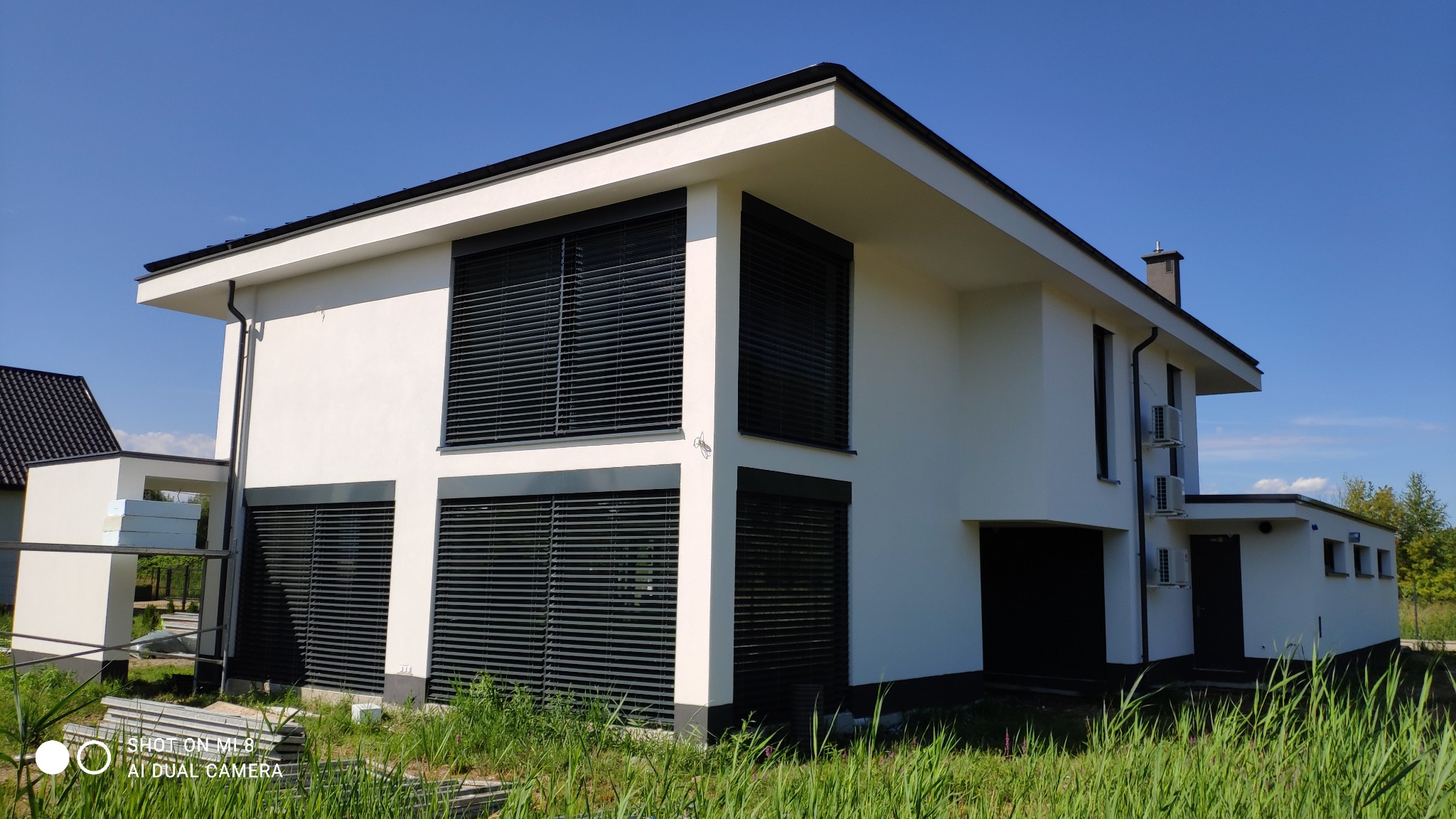 Nowoczesny, dwukondygnacyjny dom z płaskim dachem, białymi ścianami i ciemnymi żaluzjami zewnętrznymi; widoczne jednostki klimatyzacyjne na elewacji bocznej oraz materiały budowlane obok budynku.