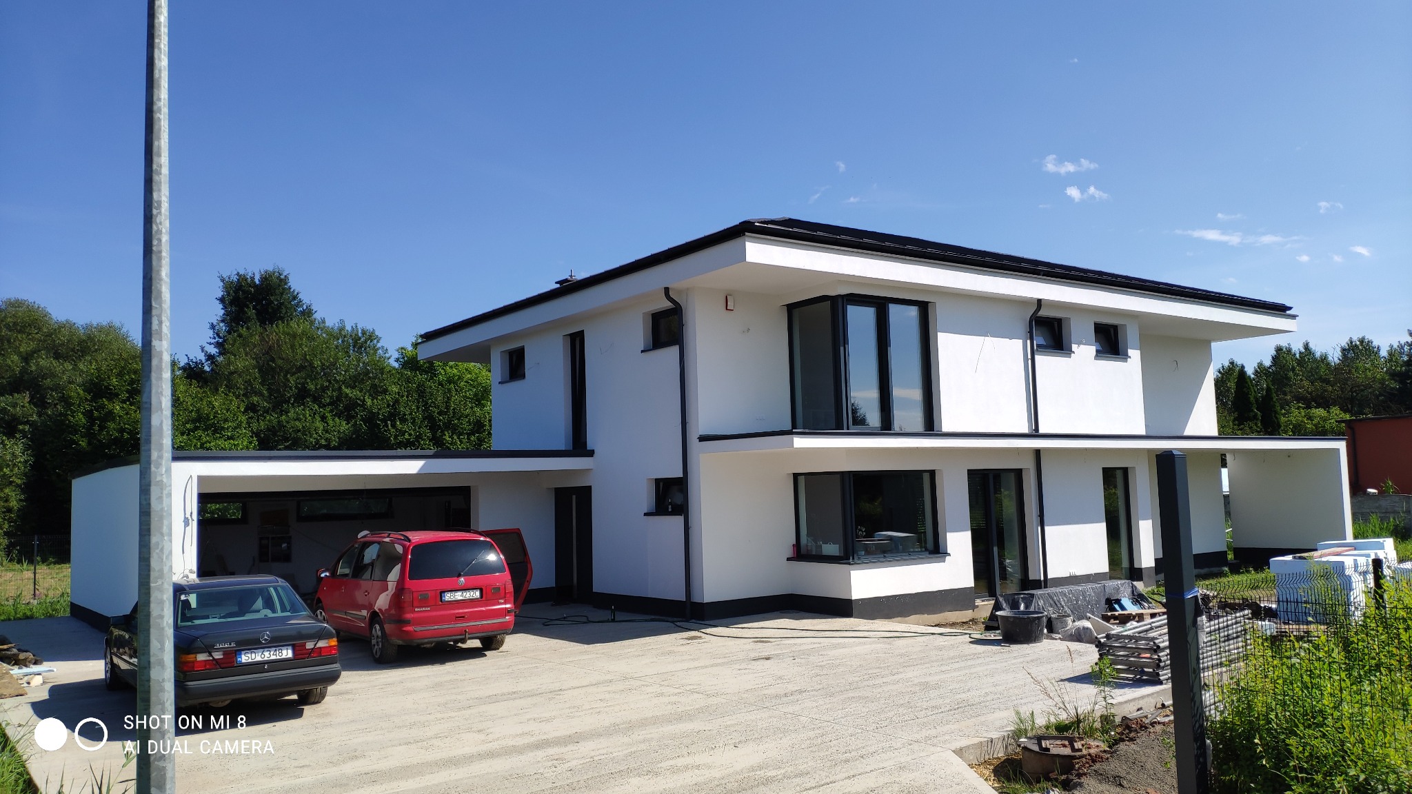 Nowoczesny, dwukondygnacyjny dom z białymi ścianami i czarnymi ramami okiennymi, widok z zewnątrz, z dwoma samochodami zaparkowanymi przed garażem, w tle zieleń drzew i błękitne niebo.