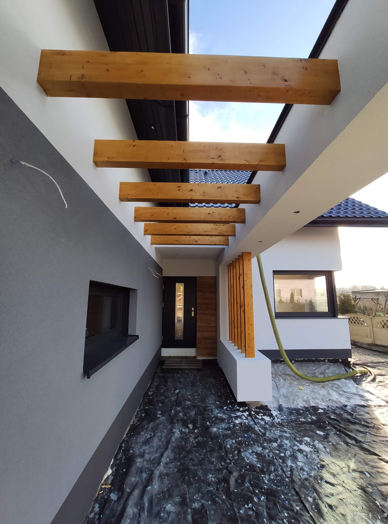 Nowoczesny dom jednorodzinny z jasnoszarą i białą elewacją, drewnianymi elementami dekoracyjnymi nad wejściem i osłoną z drewnianych listew, widoczna folia ochronna na podłożu sugerująca prace...