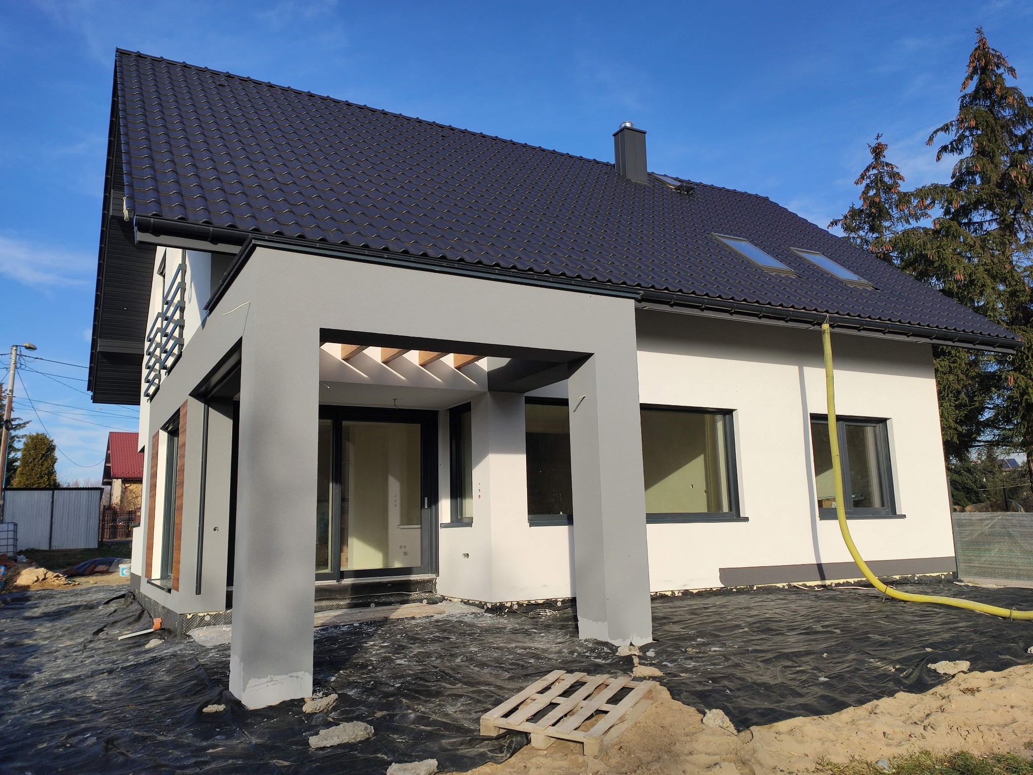 Nowoczesny dom jednorodzinny z szarą elewacją i ciemnym dachem, widoczny na etapie wykończeniowym, z rozłożoną czarną folią ochronną wokół budynku i żółtym wężem do pompowania.