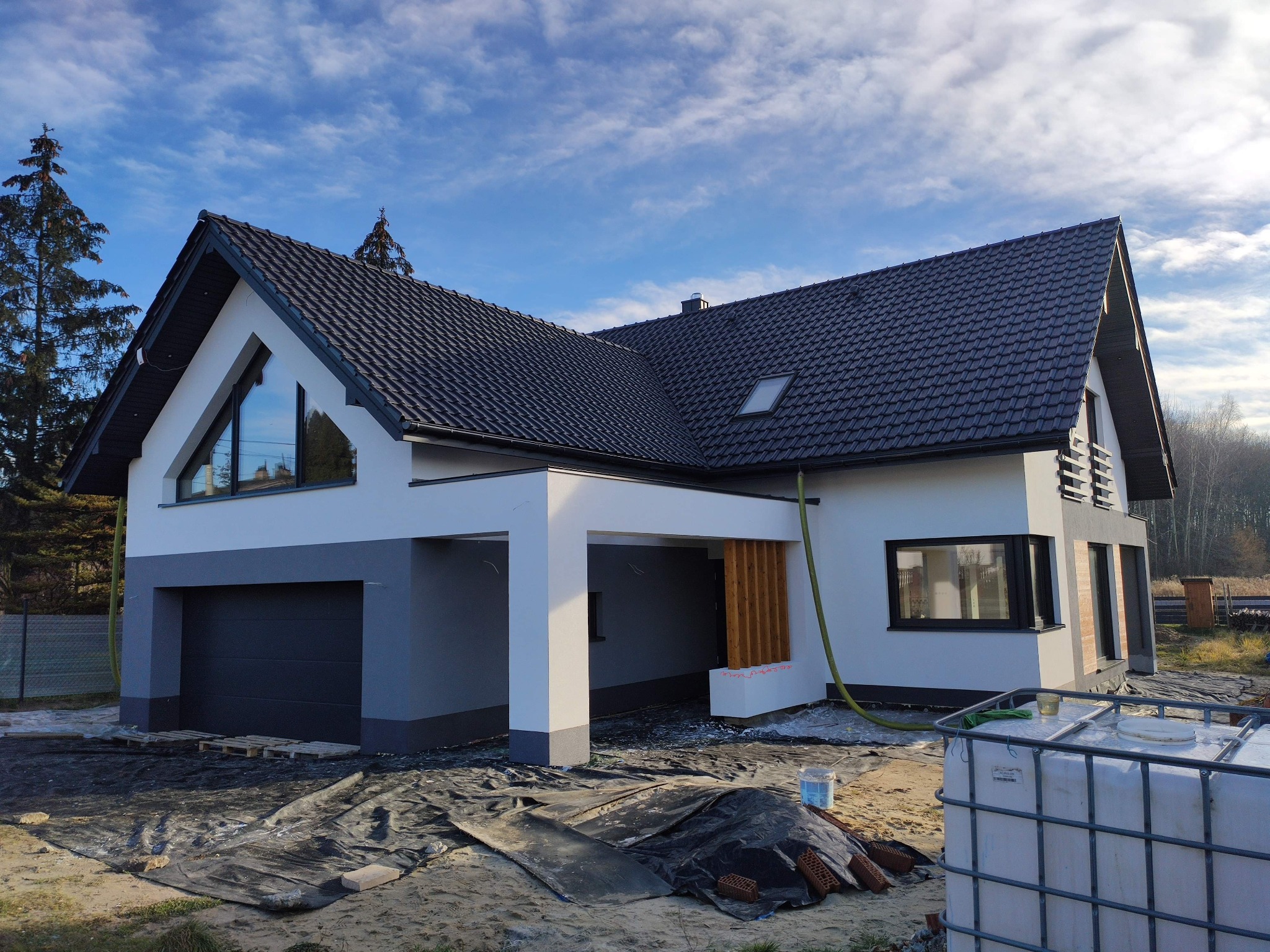 Nowoczesny dom jednorodzinny z szarą stolarką okienną i garażem w trakcie prac wykończeniowych elewacji, widoczne materiały budowlane i zbiornik na wodę.