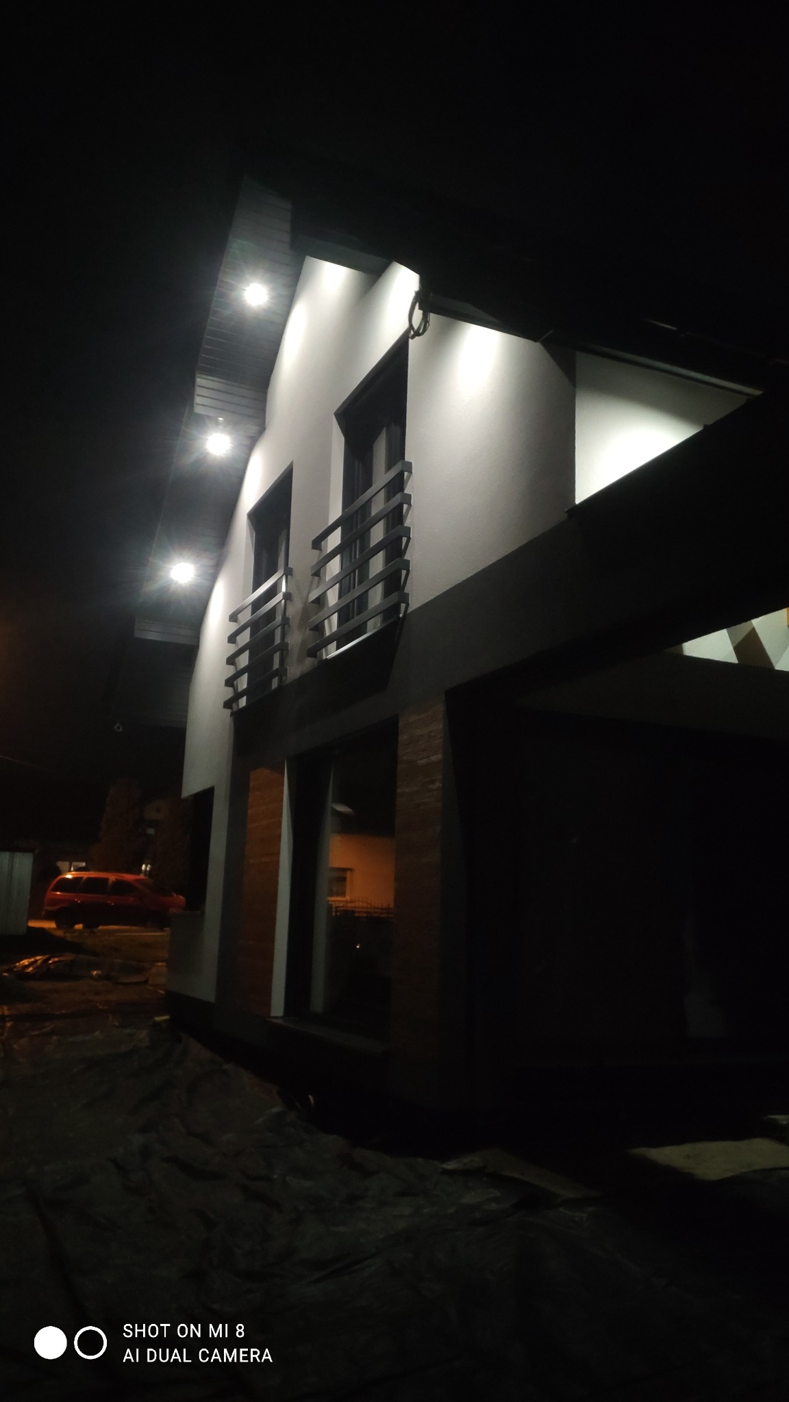 Nowoczesny dom jednorodzinny z jasną elewacją oświetloną punktowymi lampami zewnętrznymi, widoczne balkony z czarnymi balustradami, przykryte podłoże w trakcie prac.