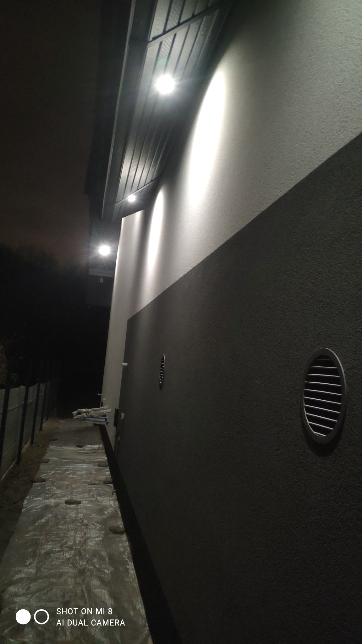 Dwukolorowa elewacja budynku oświetlona reflektorami w nocy, widoczne kratki wentylacyjne i folia ochronna na ziemi.