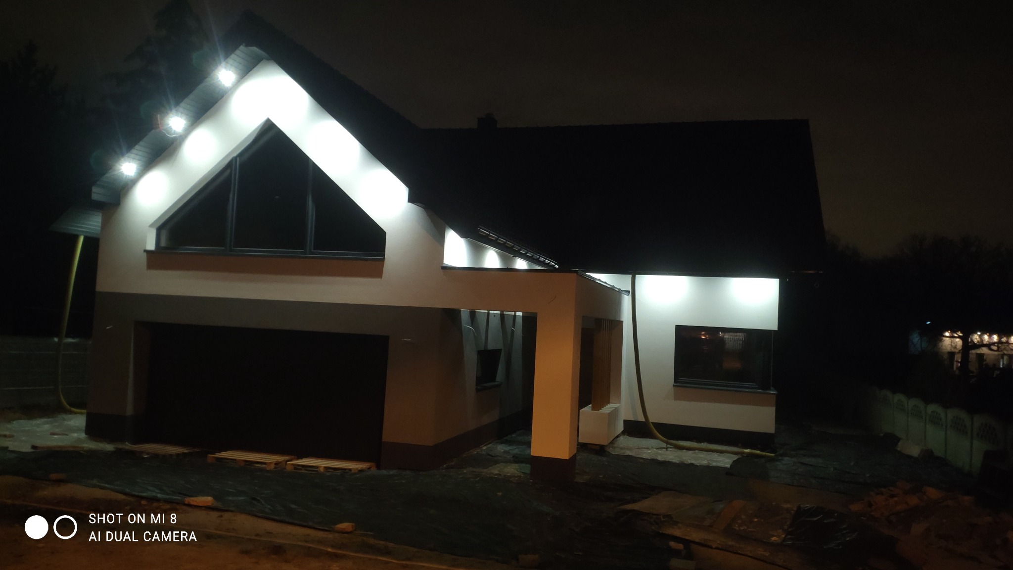 Nowoczesny dom jednorodzinny w trakcie wykańczania elewacji zewnętrznej, oświetlony nocą punktowymi lampami LED, z widocznymi elementami zabezpieczającymi teren budowy.