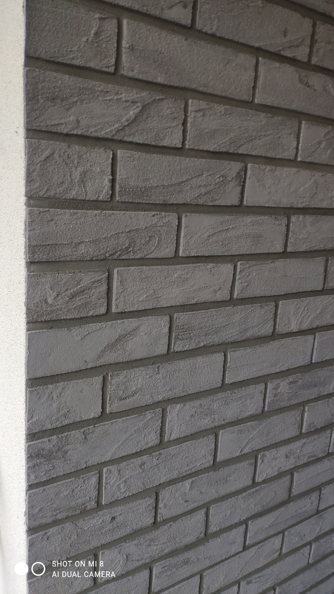 Szara elewacja imitująca cegłę, z widocznym narożnikiem budynku i delikatną fakturą powierzchni.