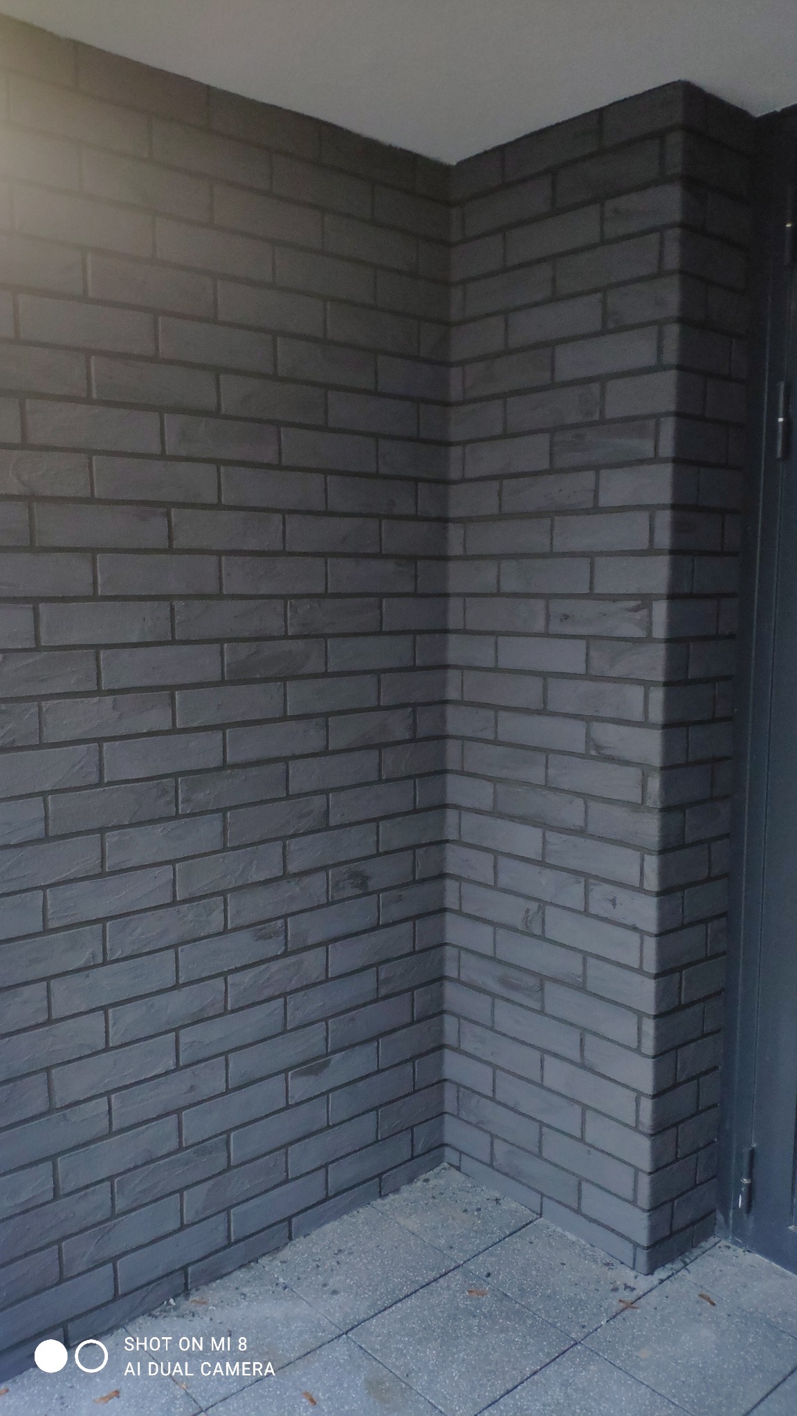 Fragment ściany zewnętrznej wykończonej szarą cegłą klinkierową z ciemną fugą, widoczny narożnik i fragment drzwi.