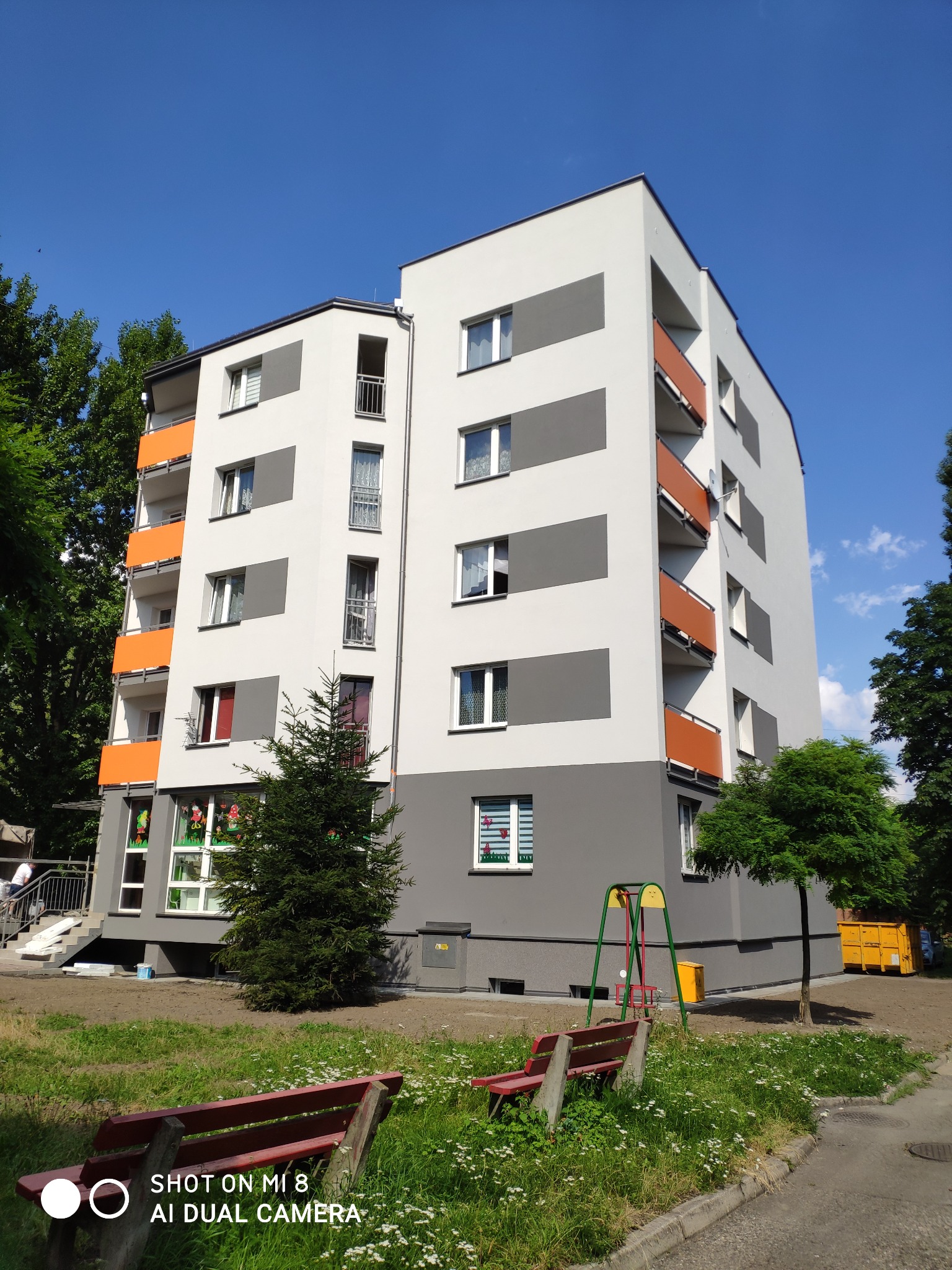 Wielopiętrowy budynek po renowacji elewacji w odcieniach szarości i bieli z pomarańczowymi balkonami, widoczny plac zabaw i zieleń.