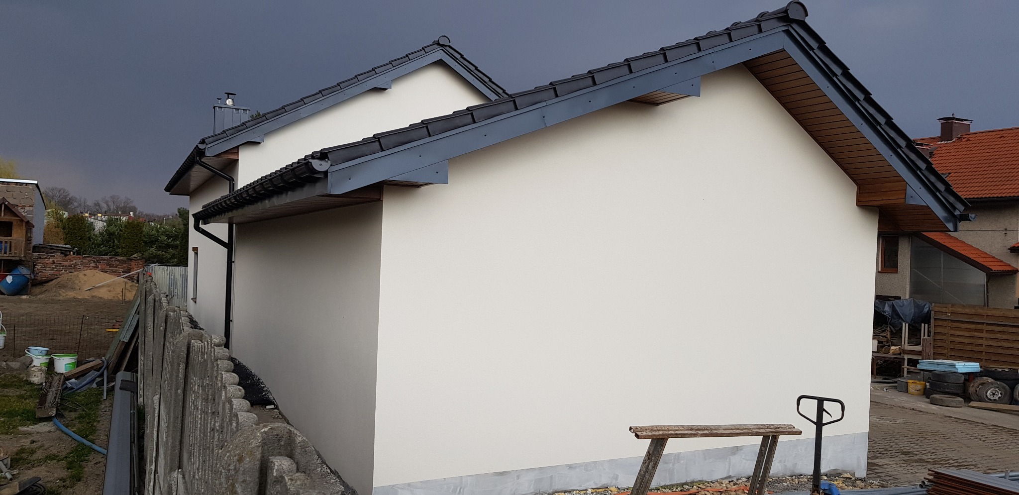 Świeżo otynkowana ściana domu jednorodzinnego w odcieniu beżu, widok z boku, ciemne dachówki i rynny, ponure niebo w tle, na pierwszym planie fragment starego, szarego płotu i prowizoryczne...